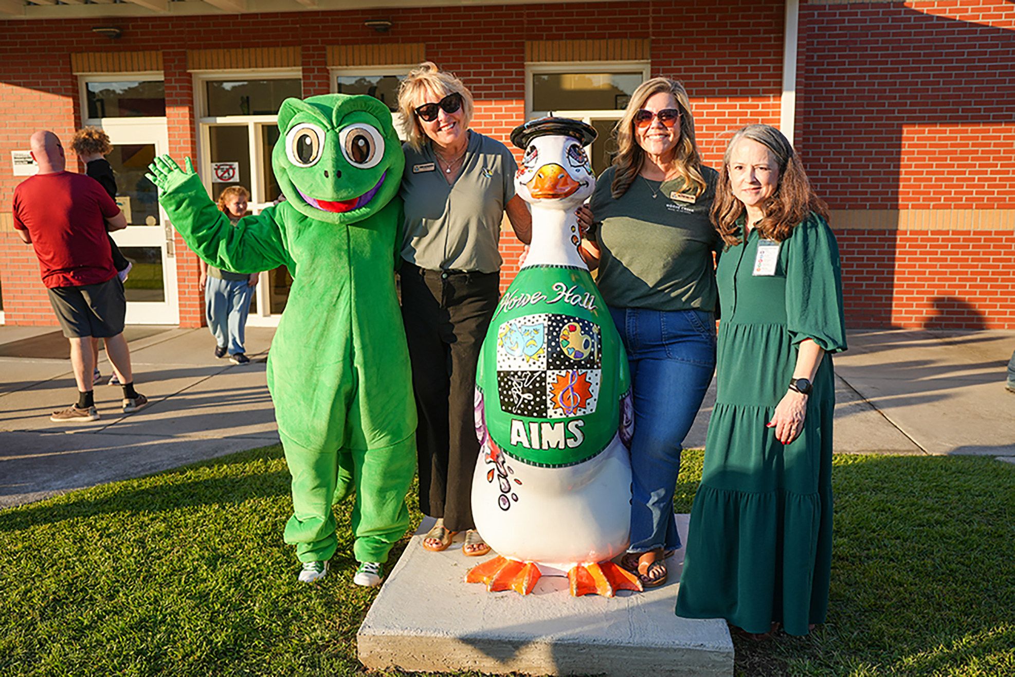 Four people in green clothing pose in front of a duck statue with "AIMS" on it, outside a brick building.