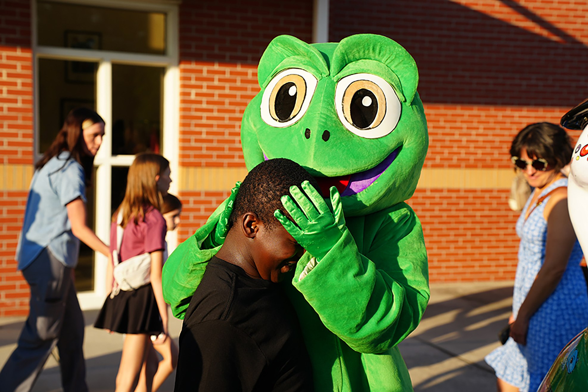 A person in a frog mascot costume hugs a young person with their hands over their face.