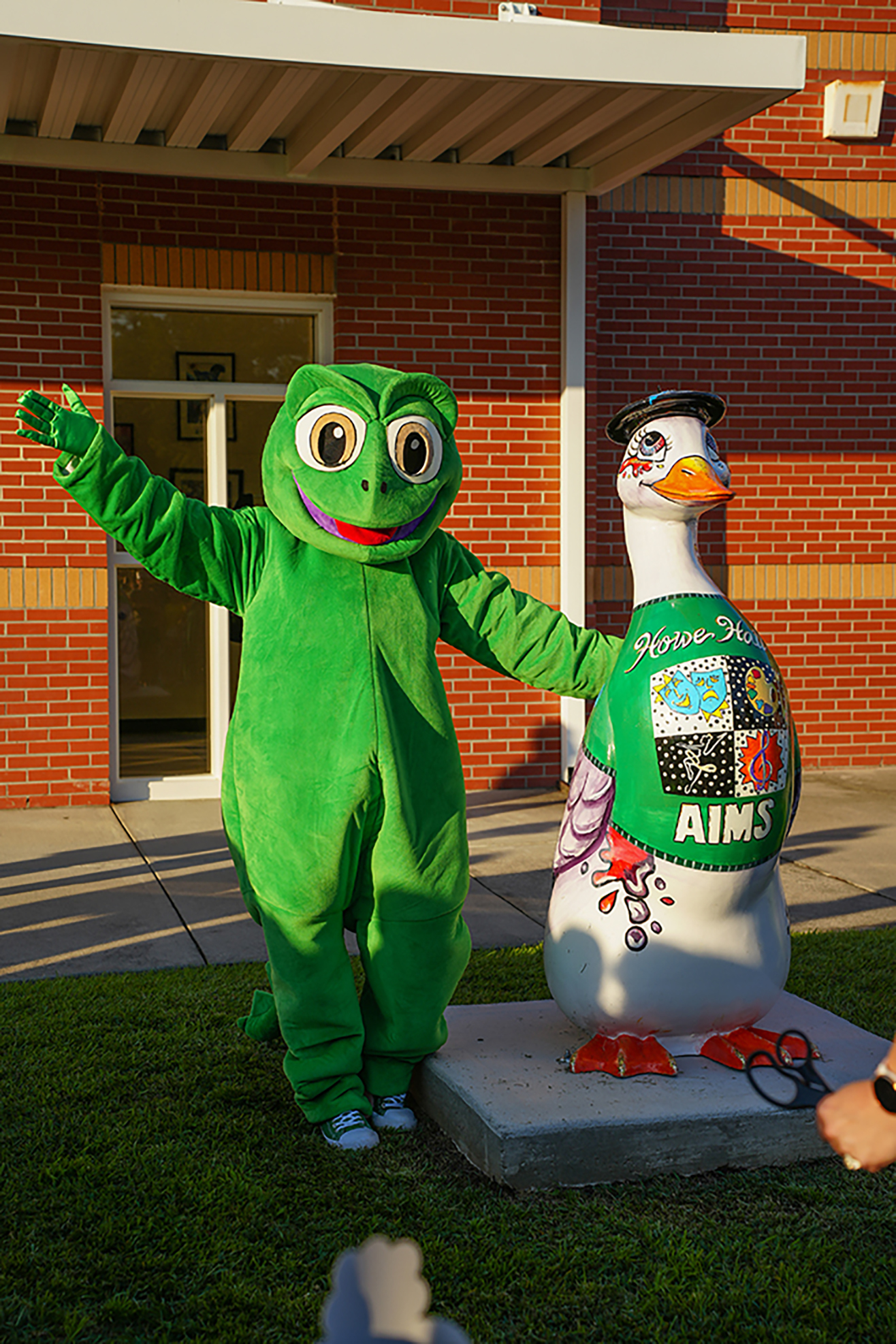 Two mascots pose in front of a brick building. One is a frog in green, the other a goose in white.