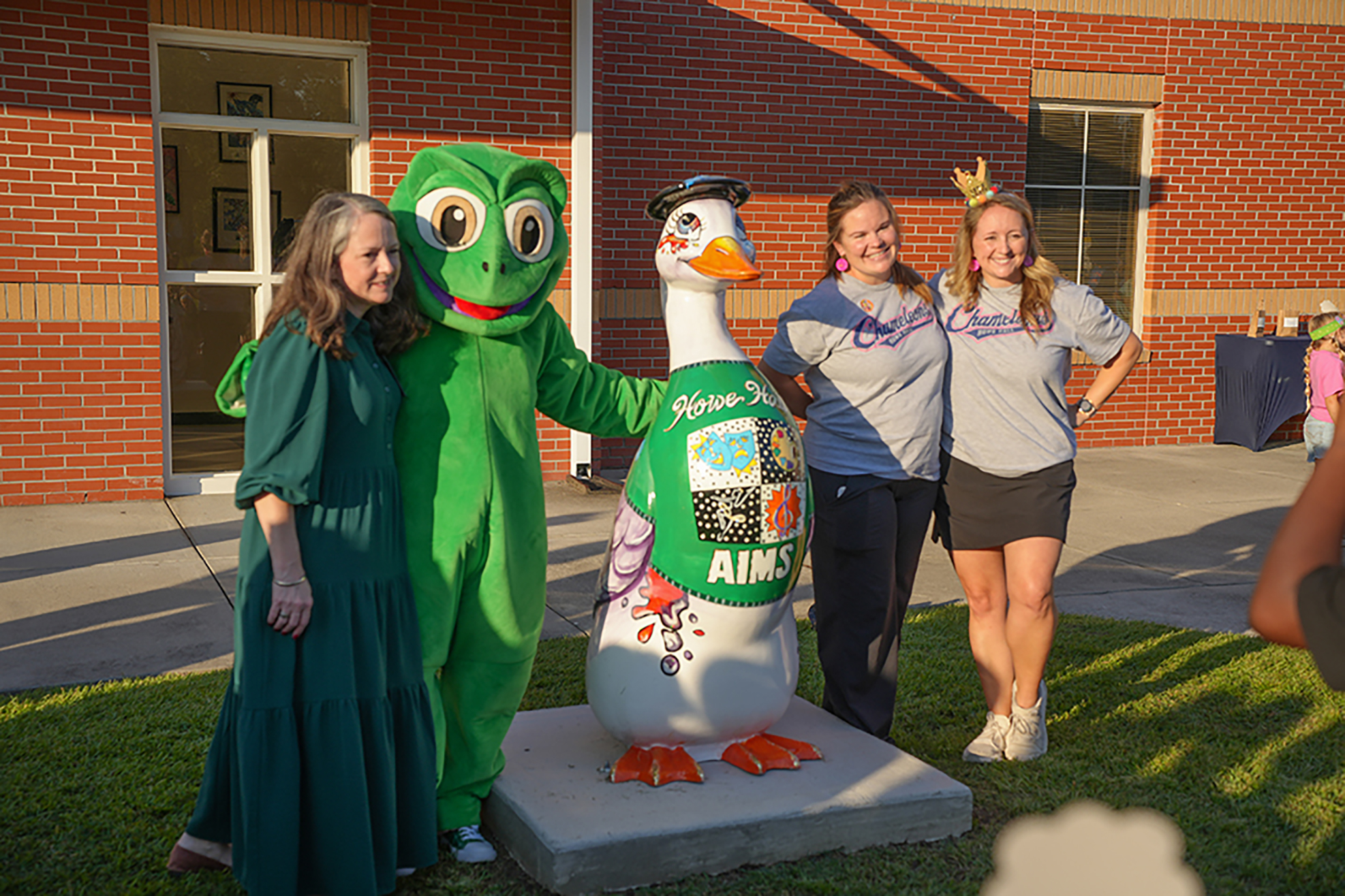 Four people, including one in a green mascot costume, stand near a brick building.