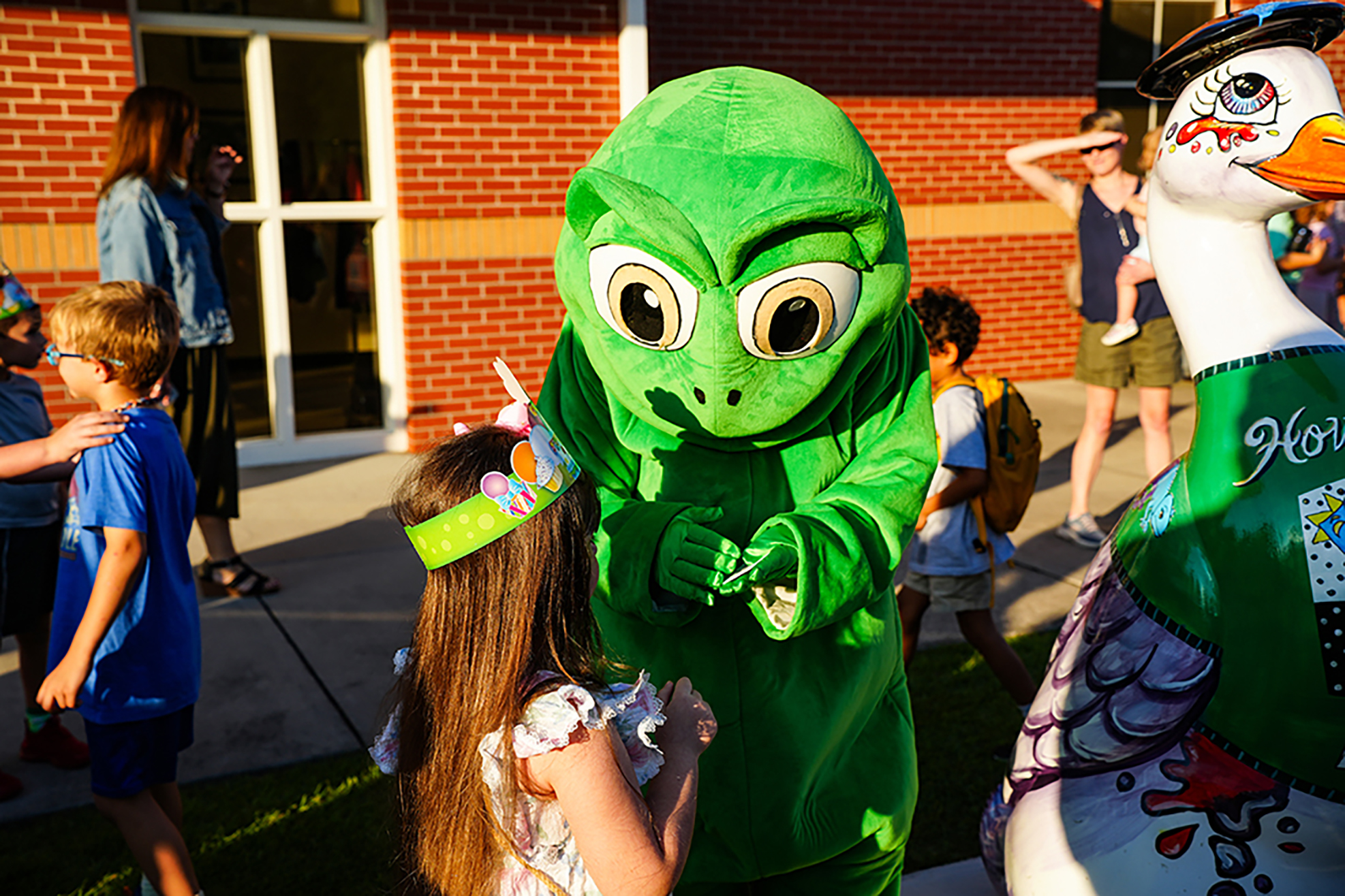 Children interact with a green mascot and a goose mascot near a brick building. Some adults observe.