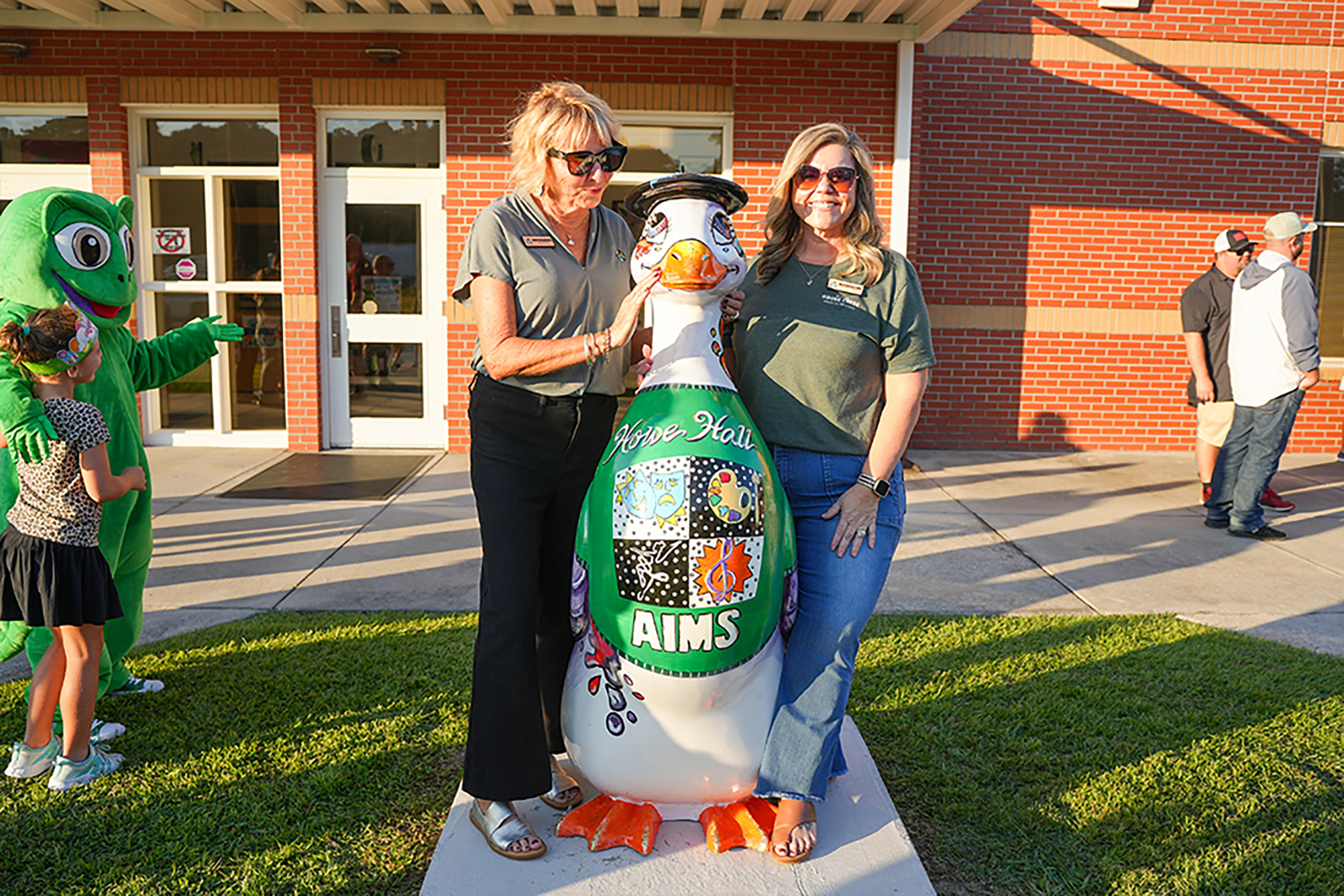 Two women in front of a building pose with a penguin statue. A green mascot is behind them.