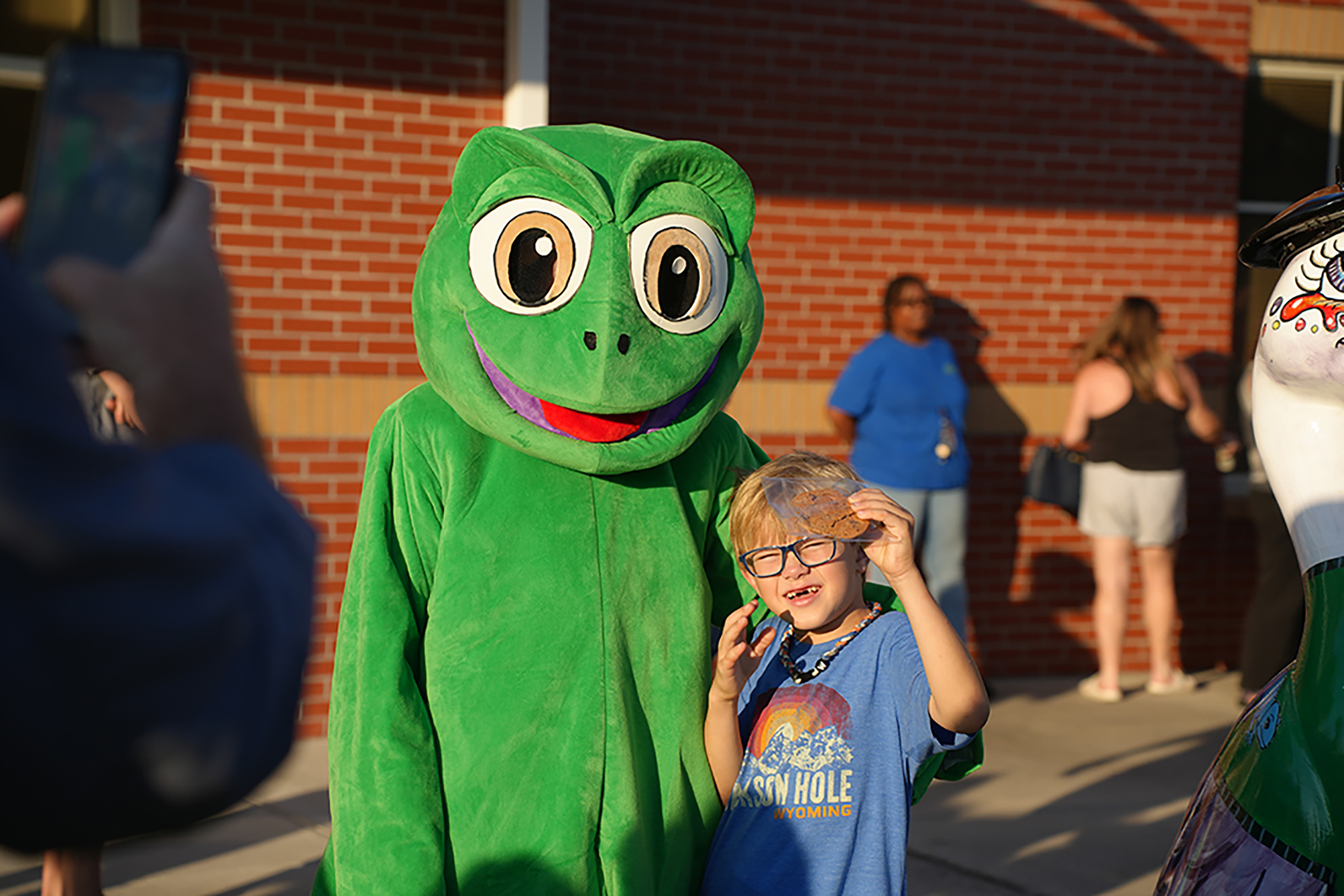A child in a blue shirt and glasses stands with a frog mascot. A woman in blue stands behind them.