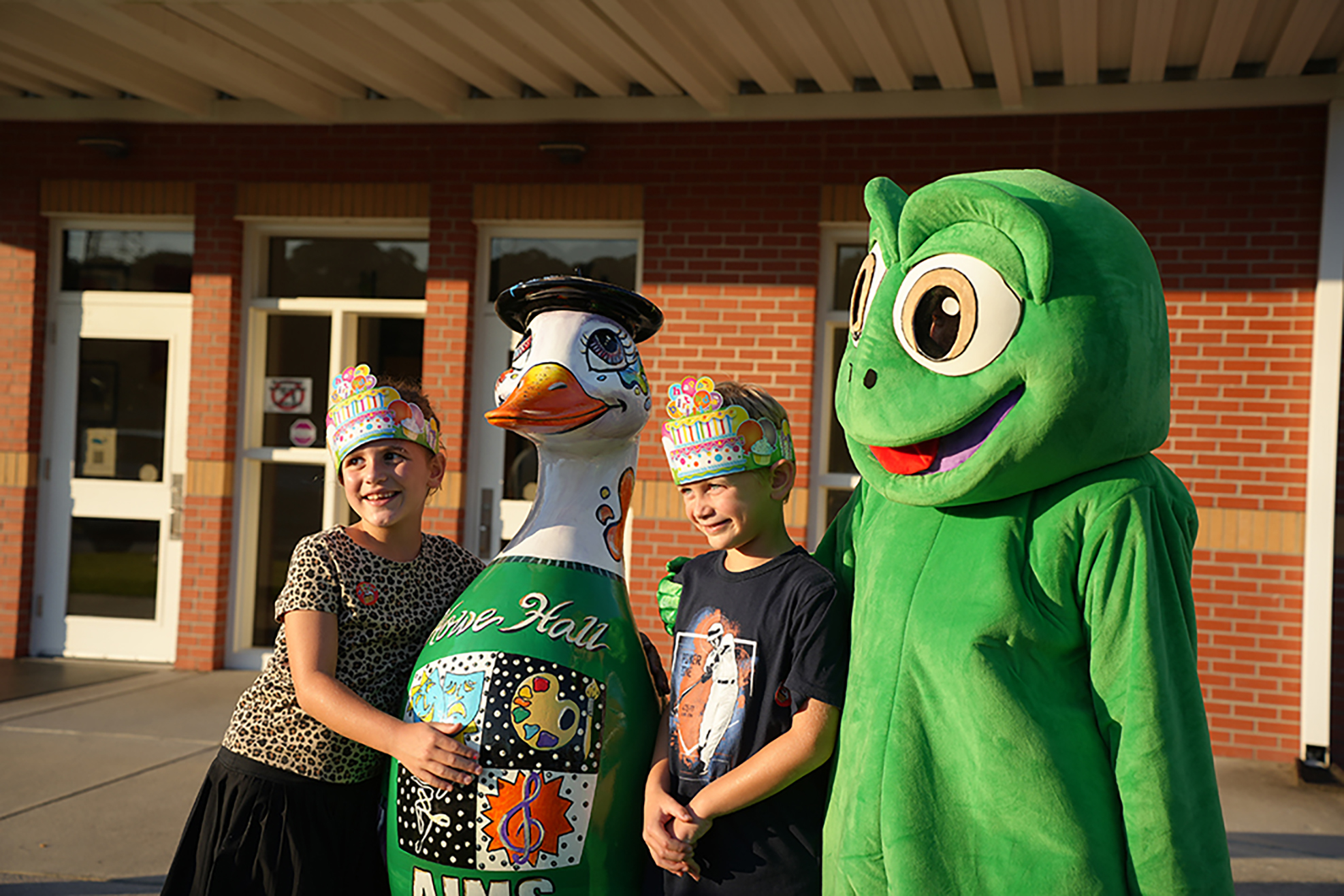 Three individuals stand in front of a brick building. Two are wearing themed hats, one is a large duck mascot, and the other is a green creature mascot.