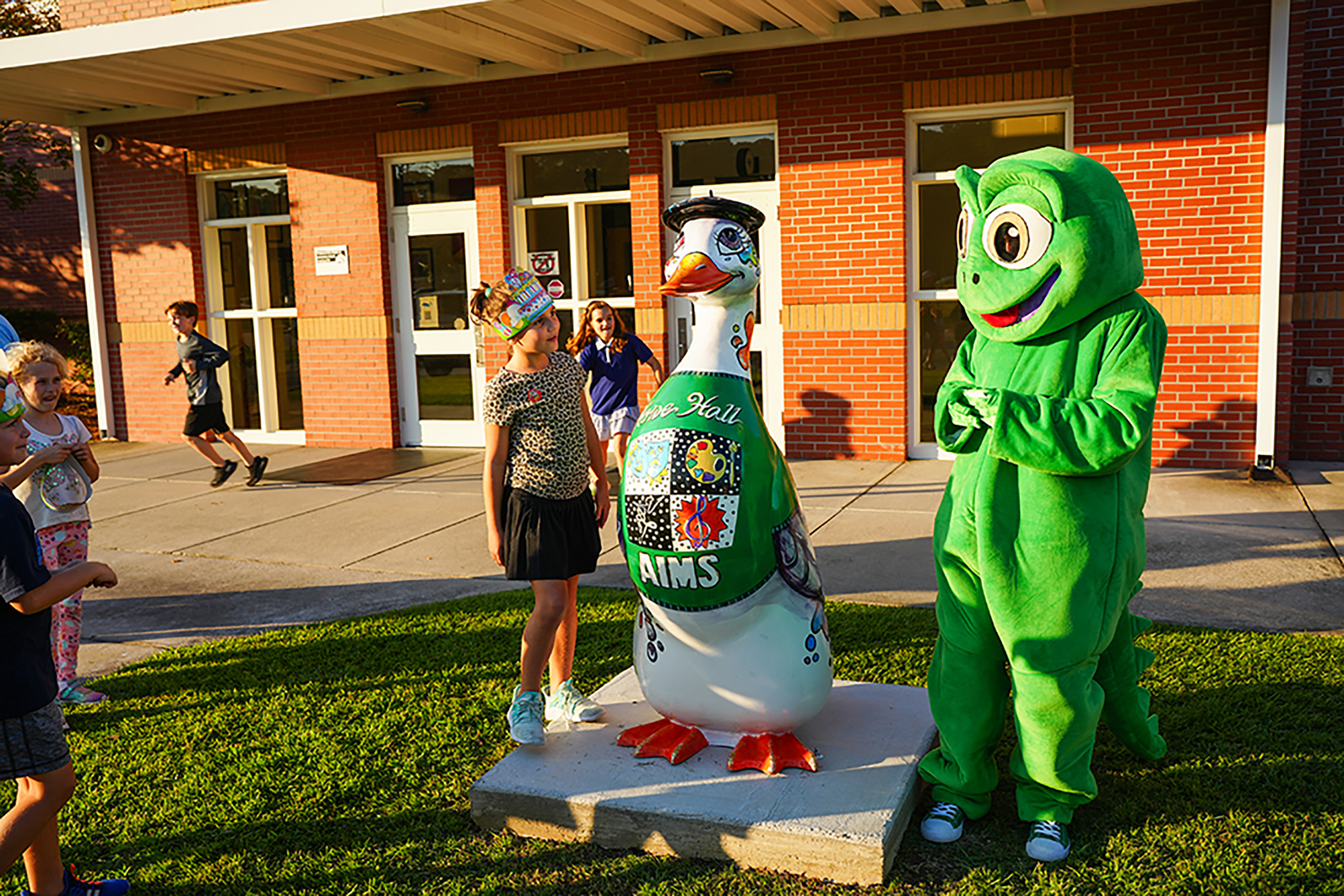 A mascot and a large goose statue on a lawn. Kids stand around them near a brick building.