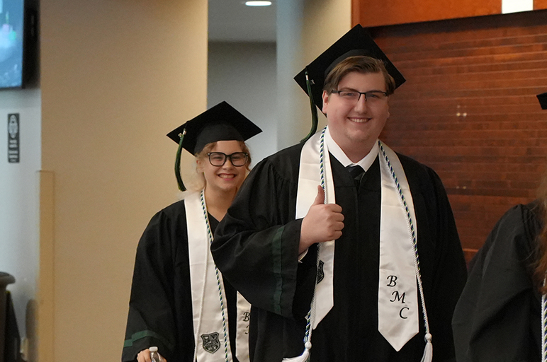 Two people in academic regalia, smiling, walking down a corridor. One wears a stole and a cap.
