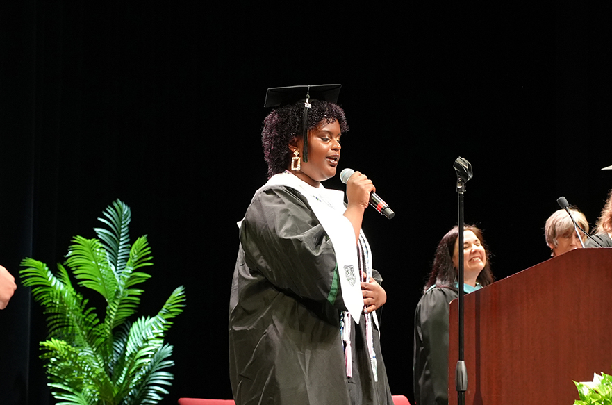Woman in cap and gown speaking into a microphone on stage; a podium, people, and a plant are behind.