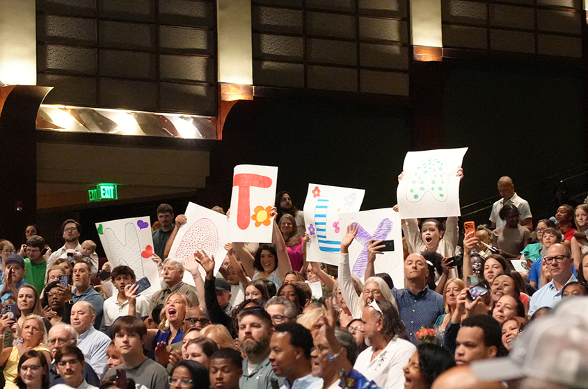 An audience of people holding signs in the air, many wearing glasses and caps. The signs have letters and designs.