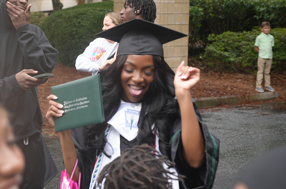 A woman in a graduation cap and gown holds a diploma, smiling and celebrating outside.