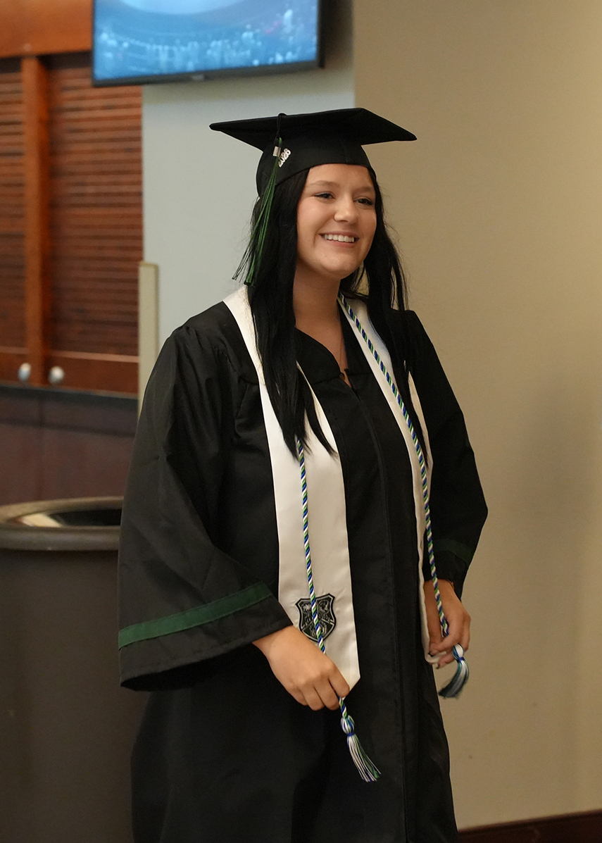 A person in a graduation gown with a mortarboard stands indoors, smiling at the camera. A trash can is to the left.