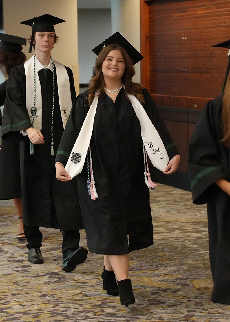 A group of individuals in black graduation gowns and caps walk in a hallway. The person in the middle wears a white sash with "BMC" and a smile.