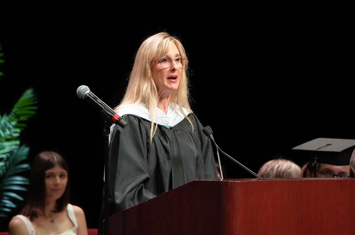A woman in a black gown stands at a podium, speaking into a microphone. Behind her, another woman watches, and others are seated.
