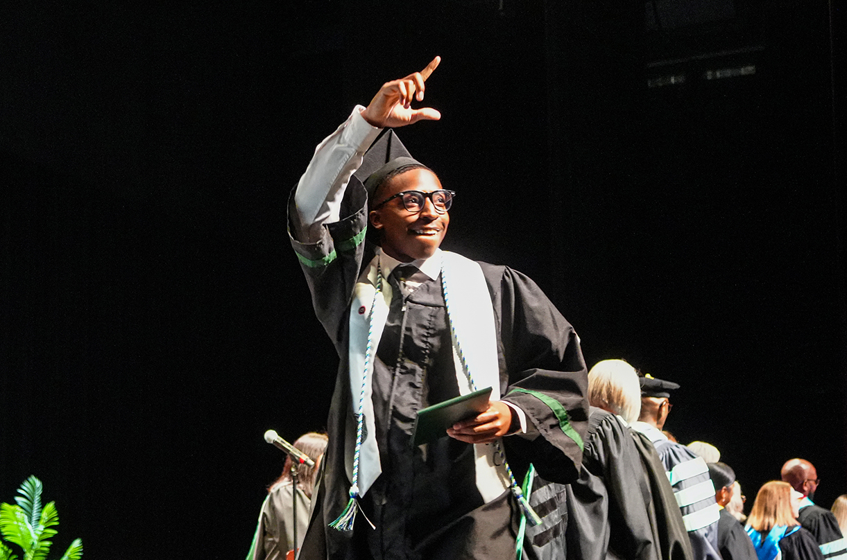A person in a graduation gown points upward on a stage. Behind them, more people in graduation robes are present.