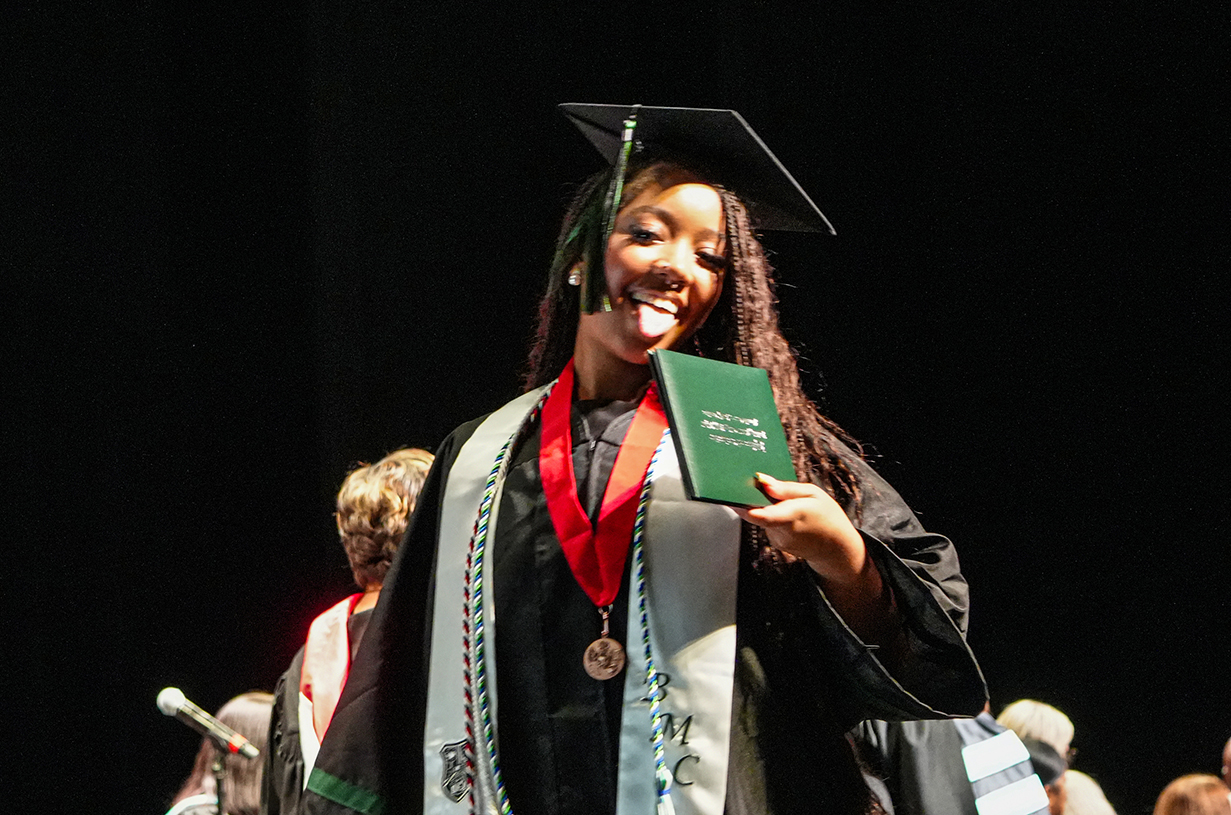 A person in a graduation gown holding a diploma smiles at the camera. Behind them, others wear gowns and hats.