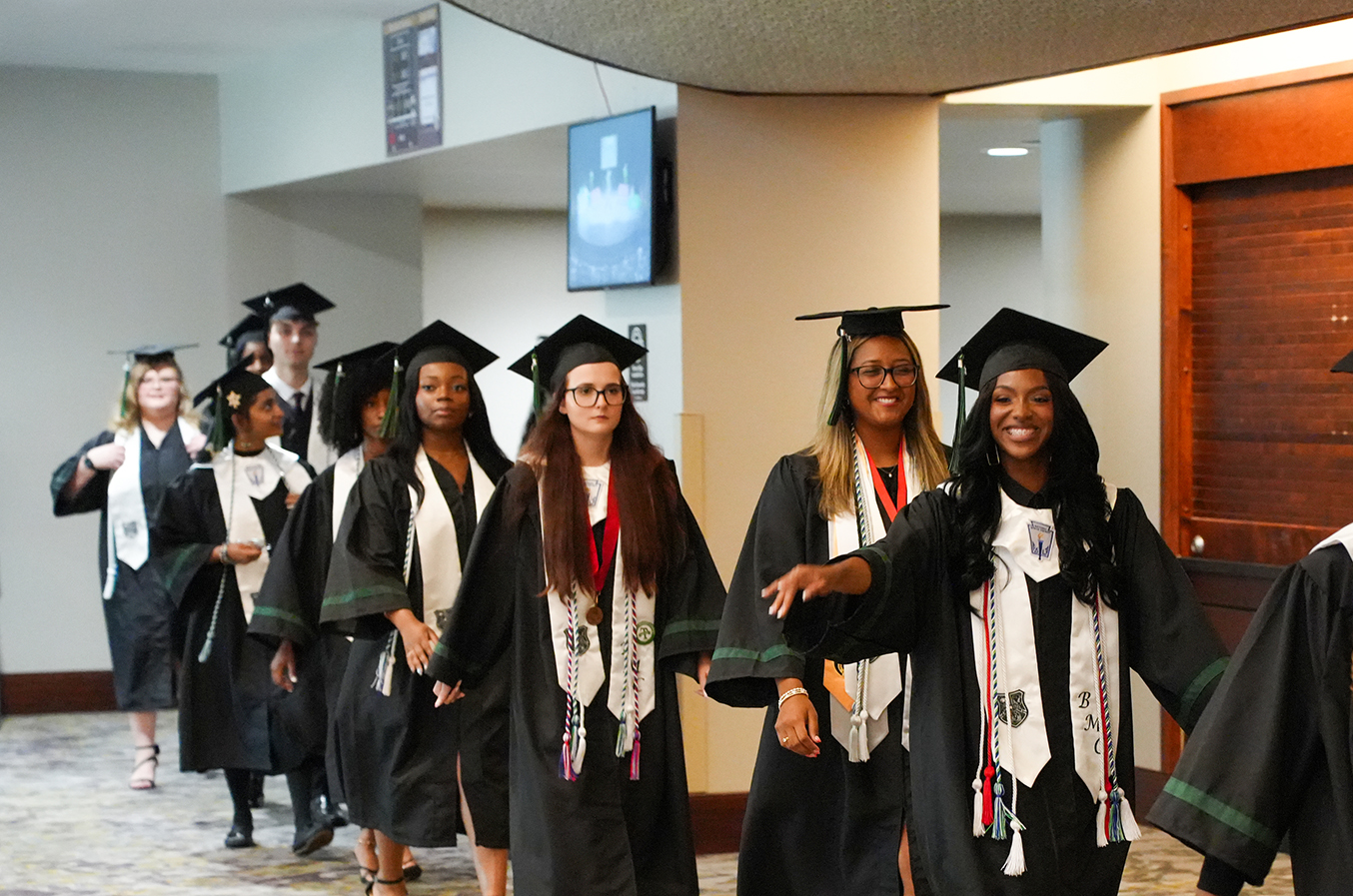 Graduates in caps and gowns walk through a hallway; some wear sashes, and one leads the group.