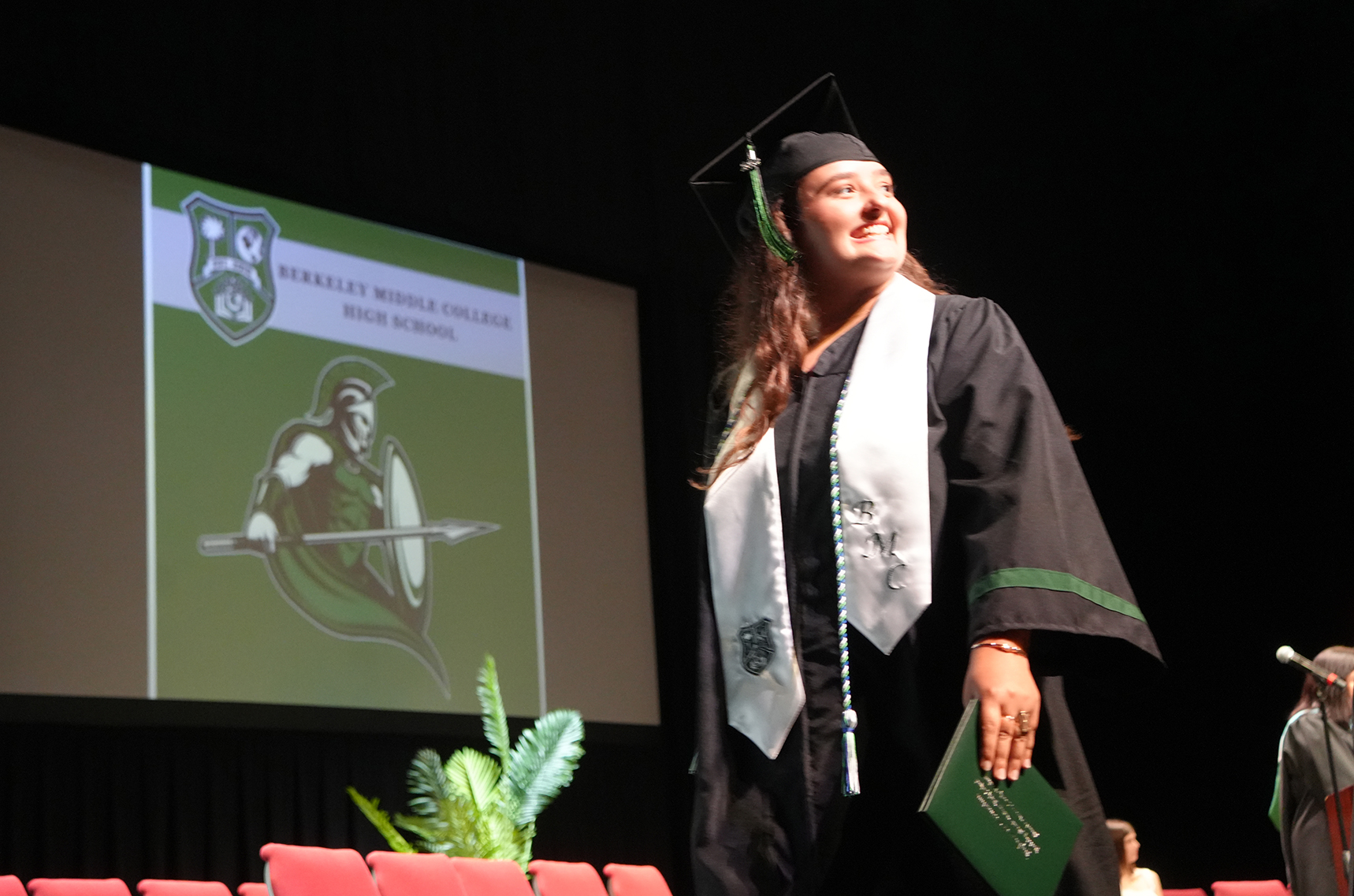 A woman in a graduation gown and cap stands on a stage with a diploma, a plant, and a large screen behind.