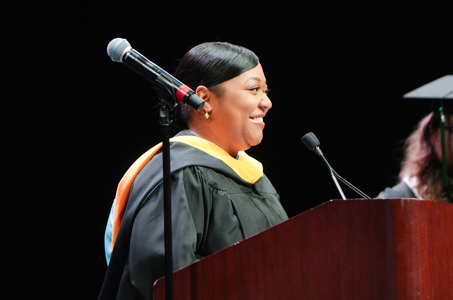 A woman in a graduation gown and stole, smiling, speaks into a microphone at a podium.