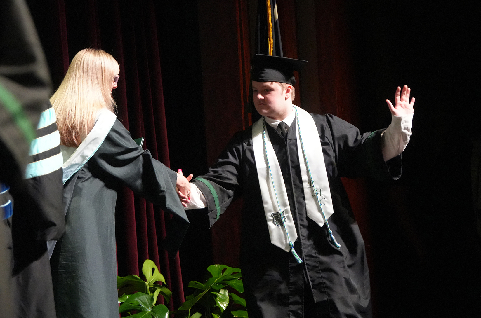 A man in a graduation gown shakes hands with a woman in a black robe on stage.