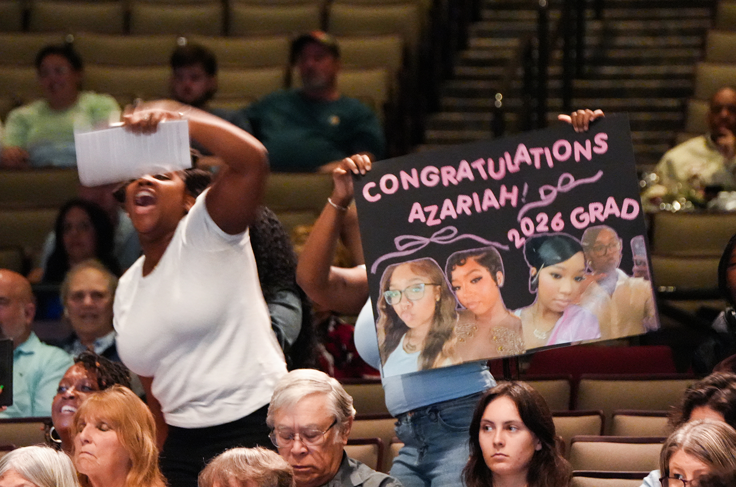 A crowd of people in an auditorium. One person holds a sign with "Congratulations Azariah, 2026 Grad" and pictures.