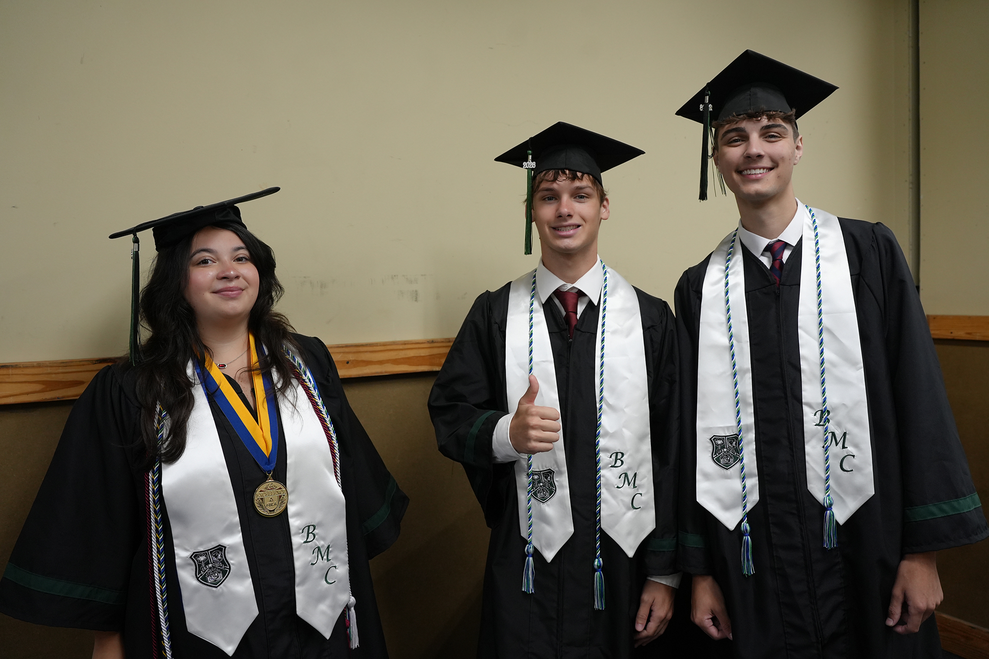 Three people in graduation attire, standing close. The woman on the left wears a medal and a smile. The man in the middle gives a thumbs-up.