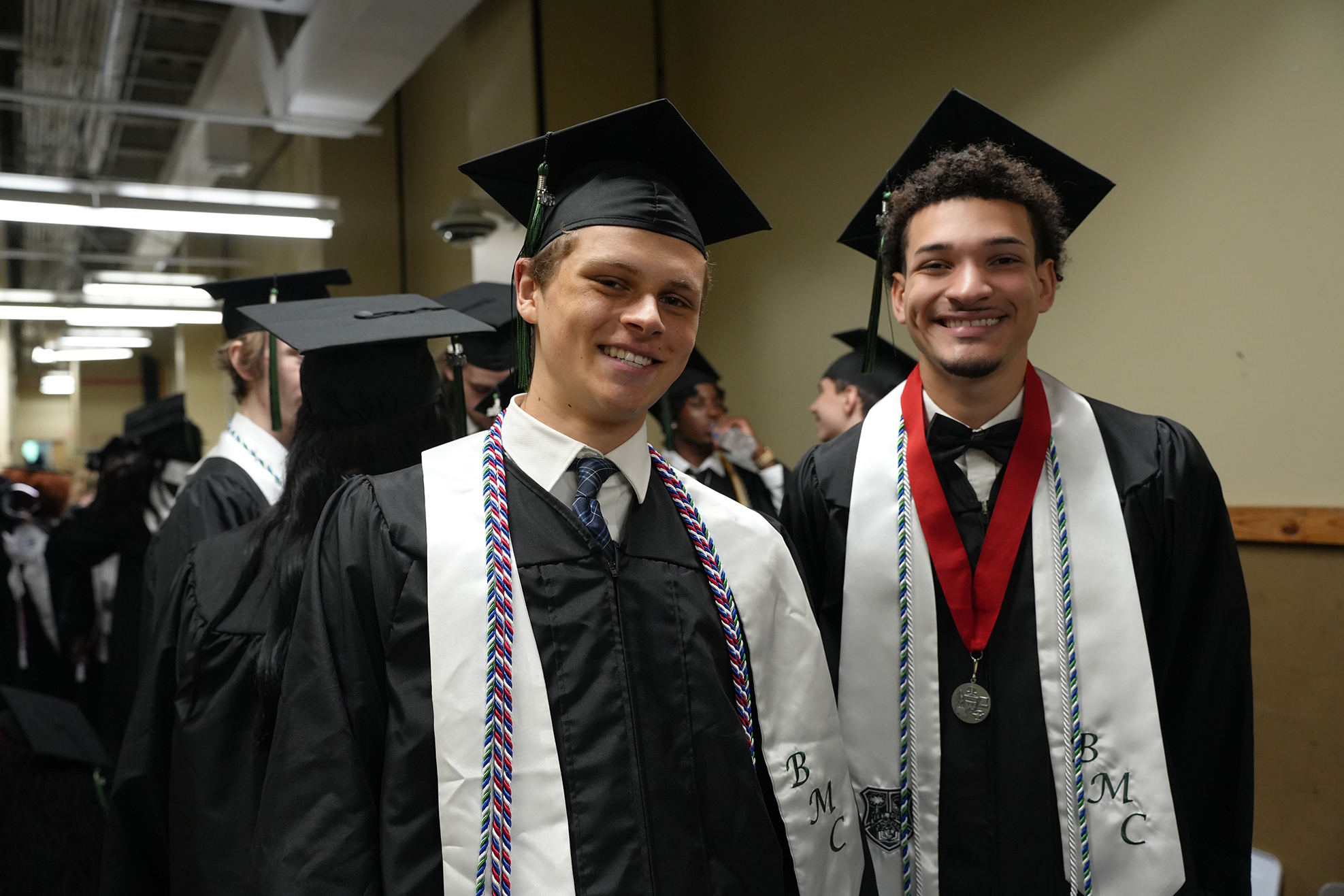 Two individuals in academic regalia stand side by side. Both wear caps and gowns, with one sporting a red bow tie.