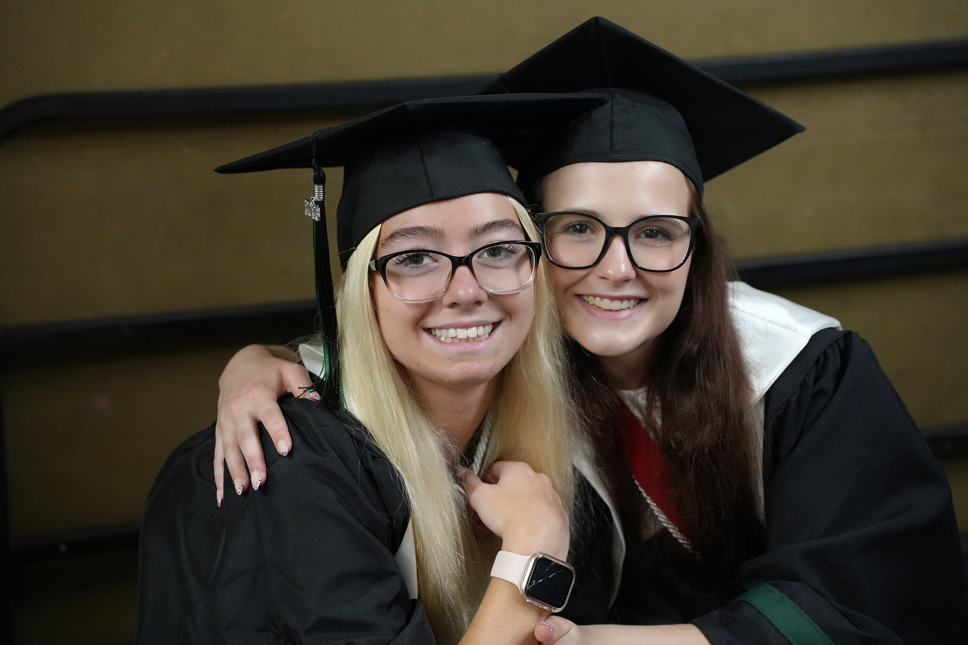 Two women in graduation gowns and caps, one with blonde hair and glasses, the other with brown hair, smiling and posing for a photo.
