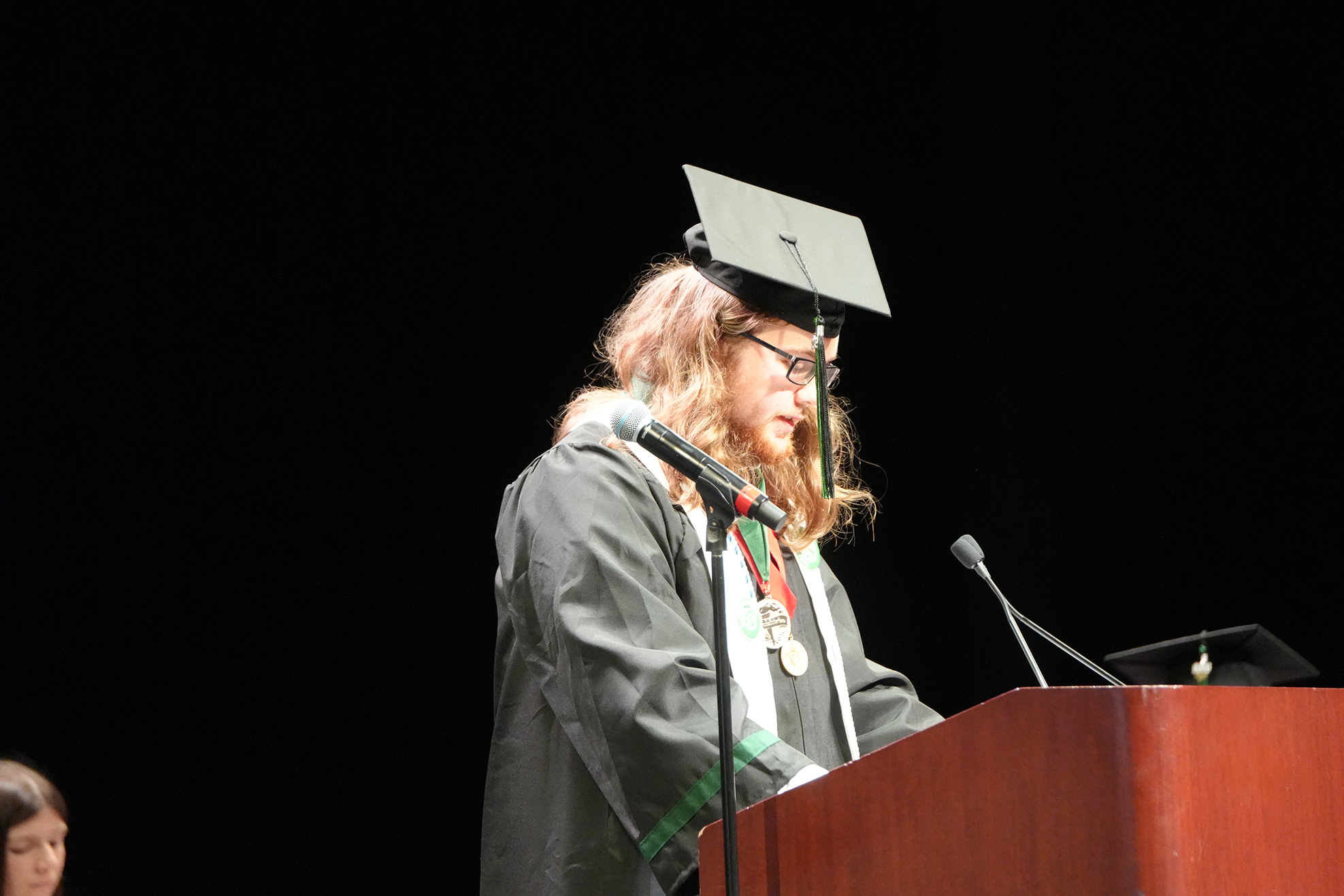 A person in a cap and gown stands at a podium with a microphone, likely speaking at an event.