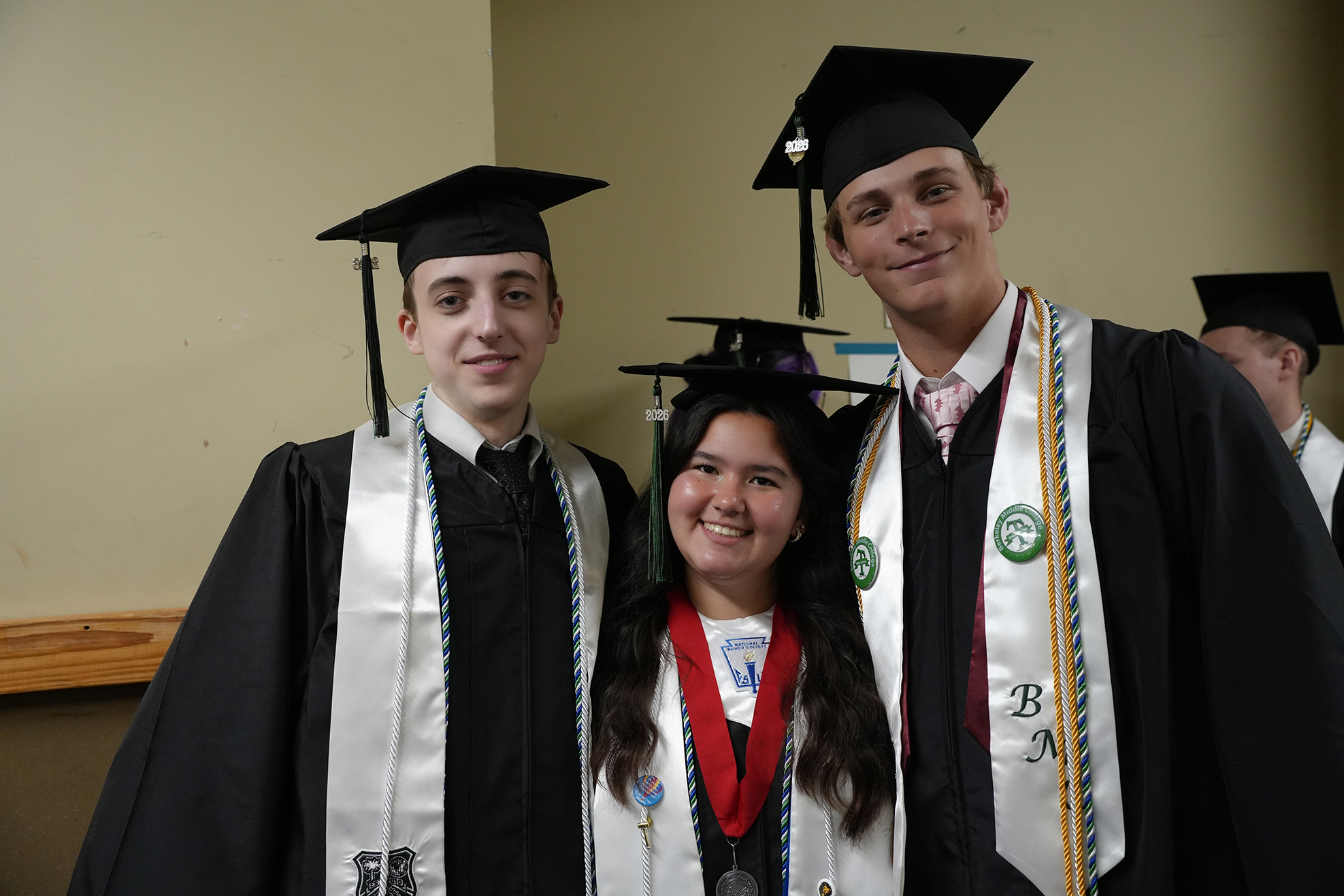 Three young adults in graduation robes and caps. The two men wear sashes. The woman wears a medal.