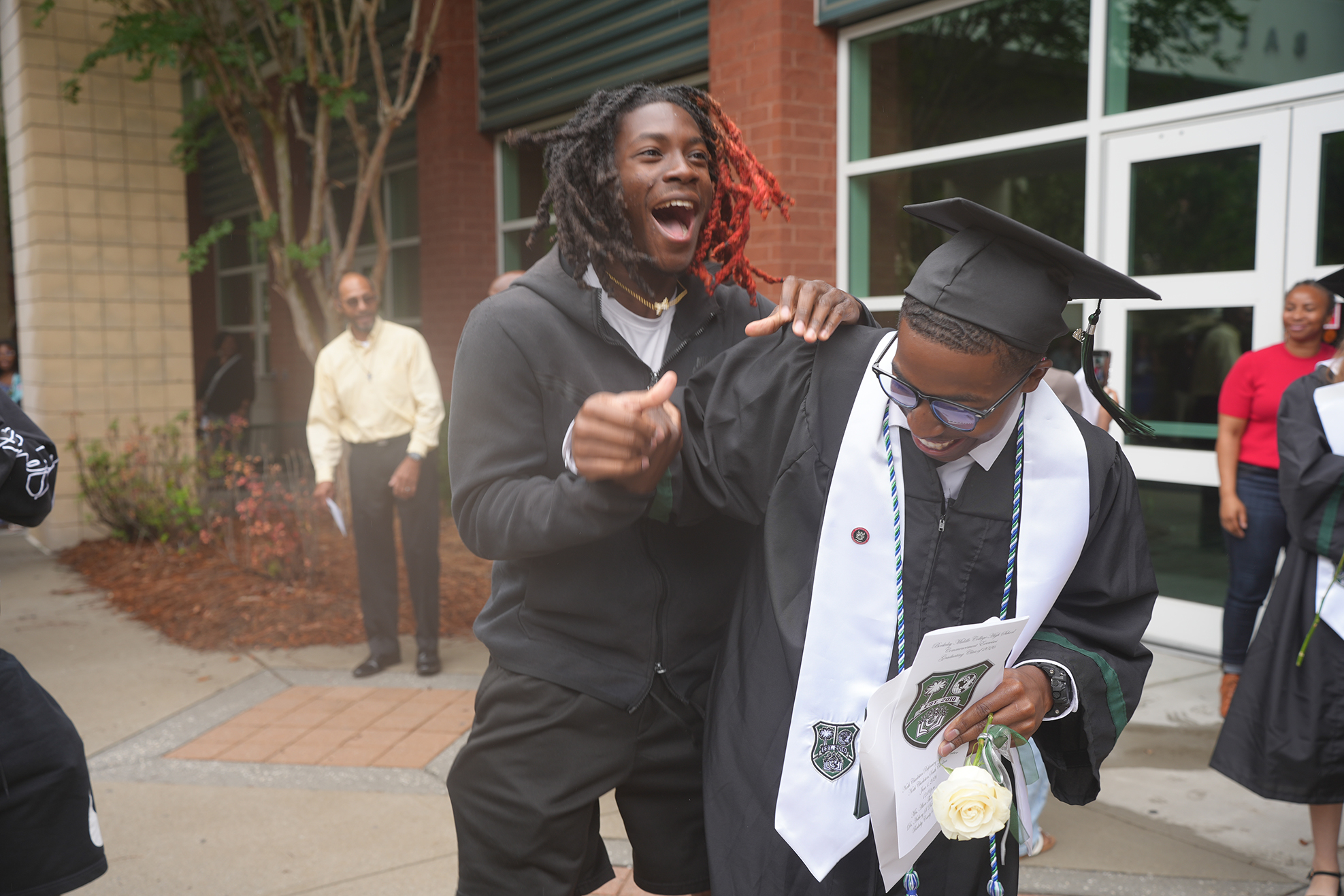 Two men in graduation attire, one in a cap and gown, the other in a hoodie, standing outdoors.