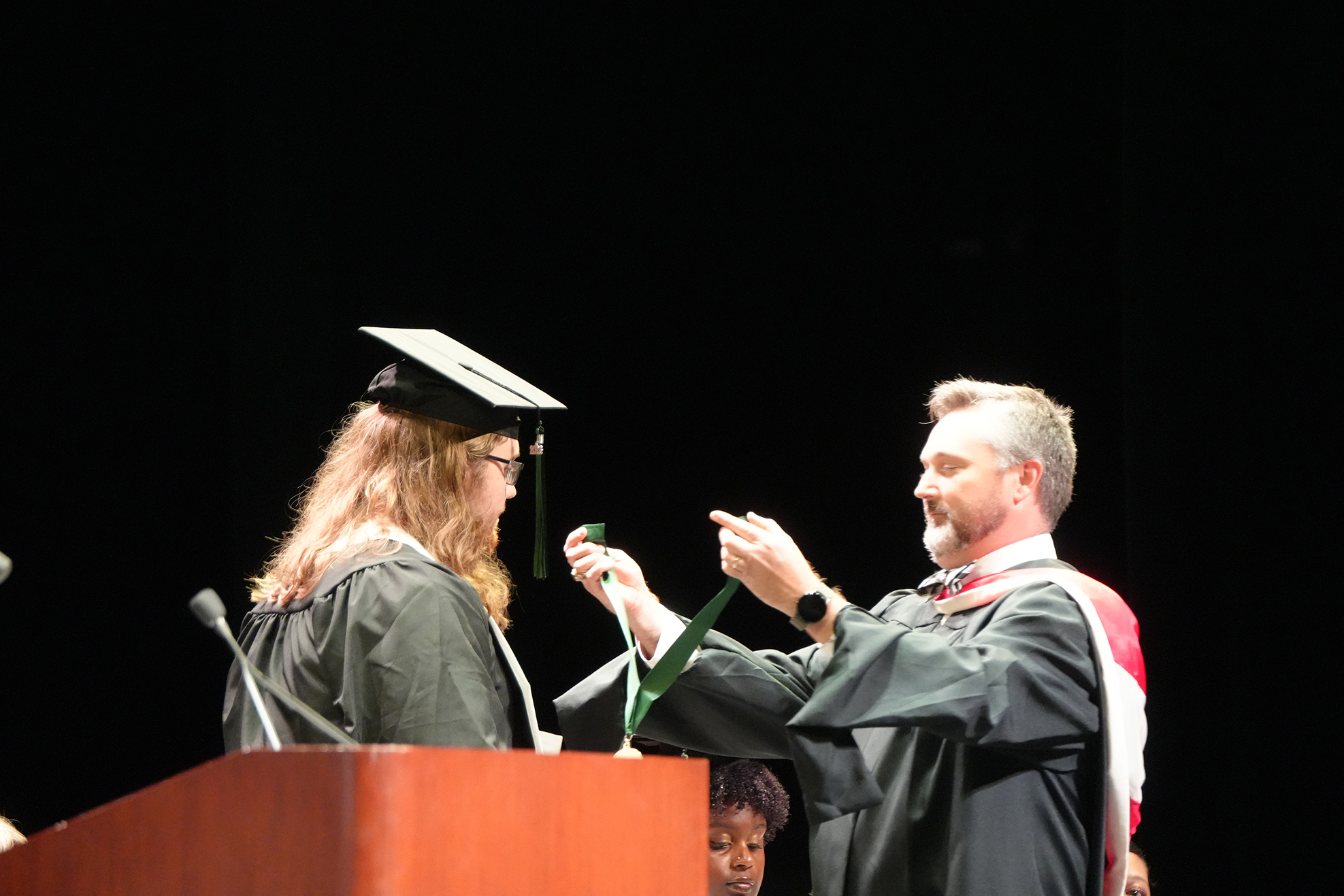 A woman in a graduation cap places a green ribbon around the neck of a man in a cap and gown.