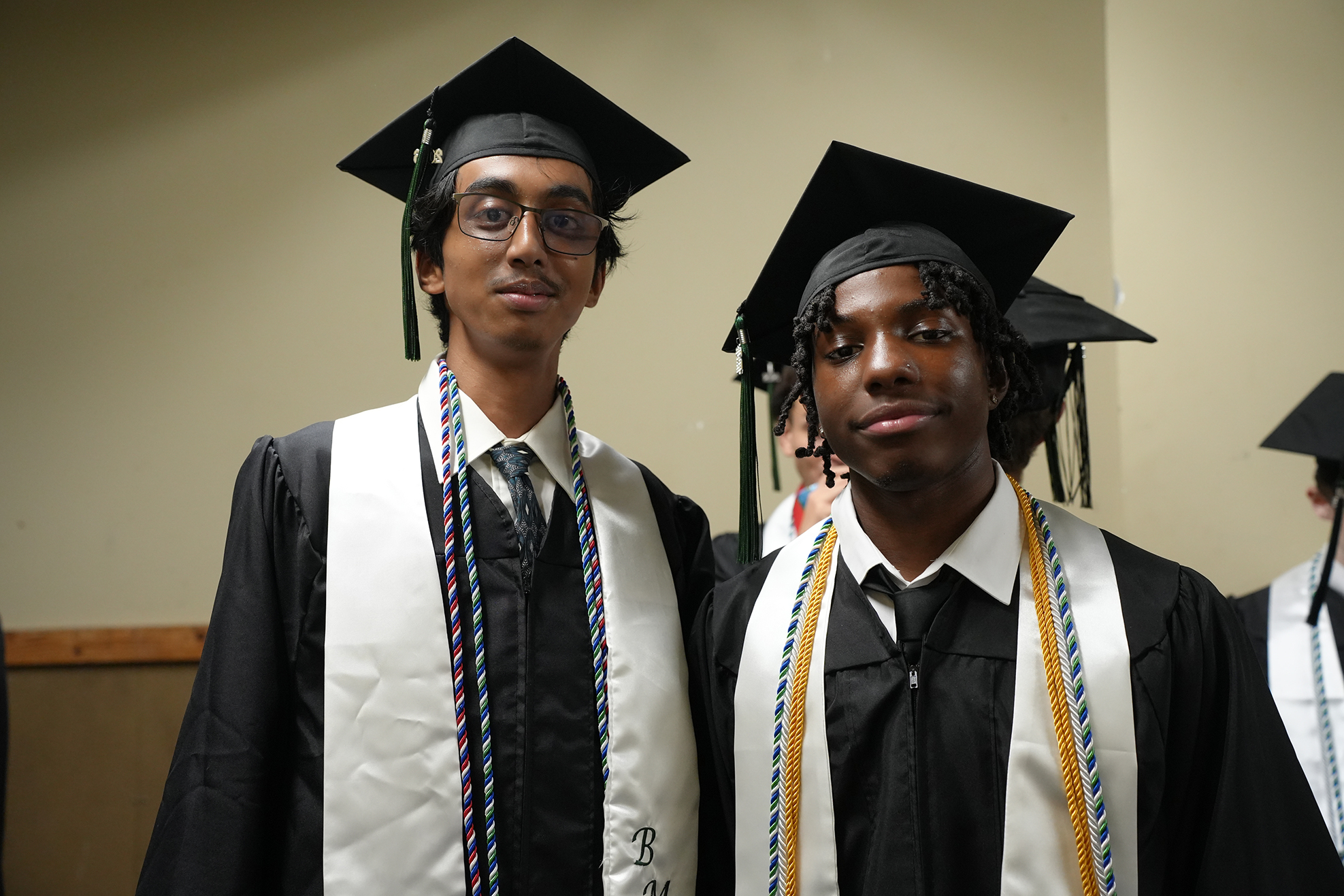 Two men in graduation robes and caps stand side by side. One has glasses and a sash with multicolored cords.