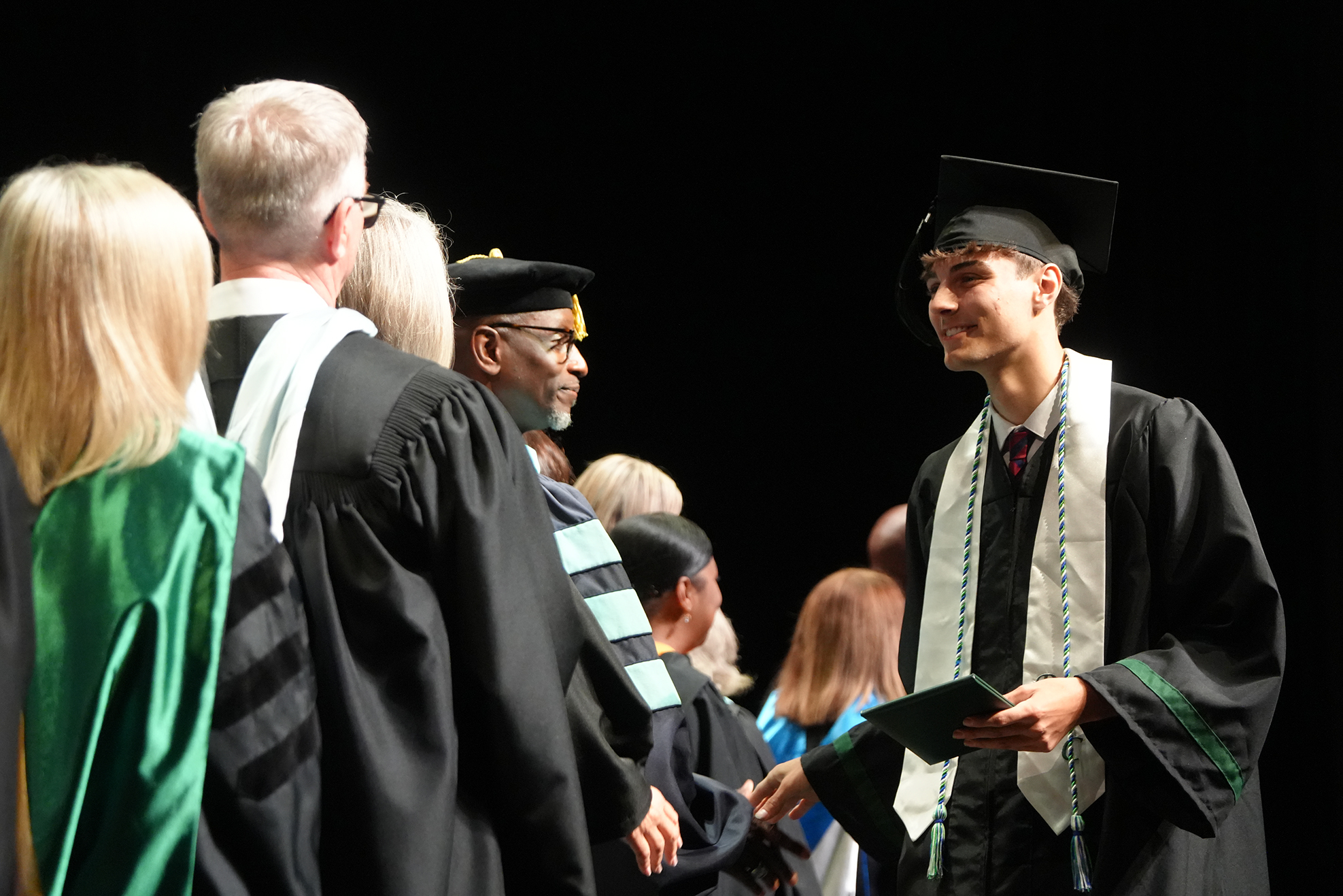 A line of individuals in academic regalia, one holding a diploma and speaking to another in a black gown.