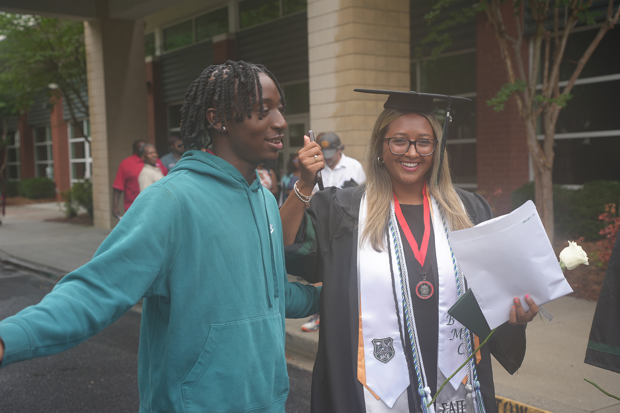 A man in a teal hoodie talks with a woman wearing a graduation cap and gown.