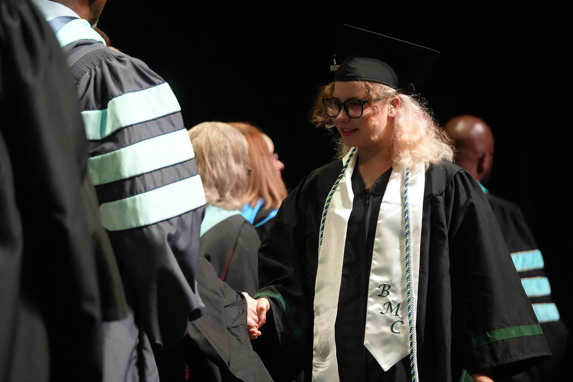 A person with blonde hair wearing a graduation cap and gown smiles while shaking hands with another person.