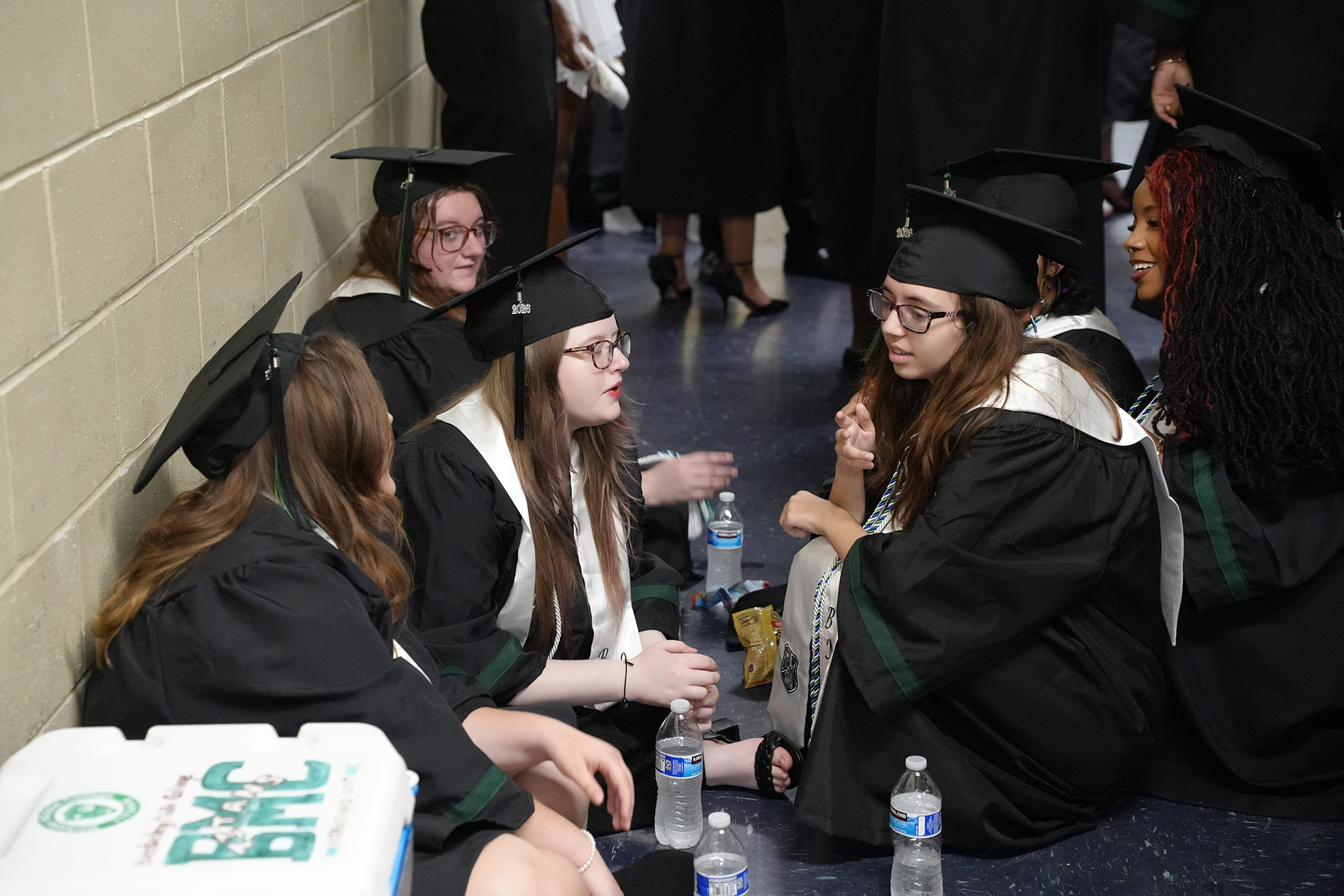 People in black graduation gowns and caps sit along a wall. Some wear glasses and carry water bottles.