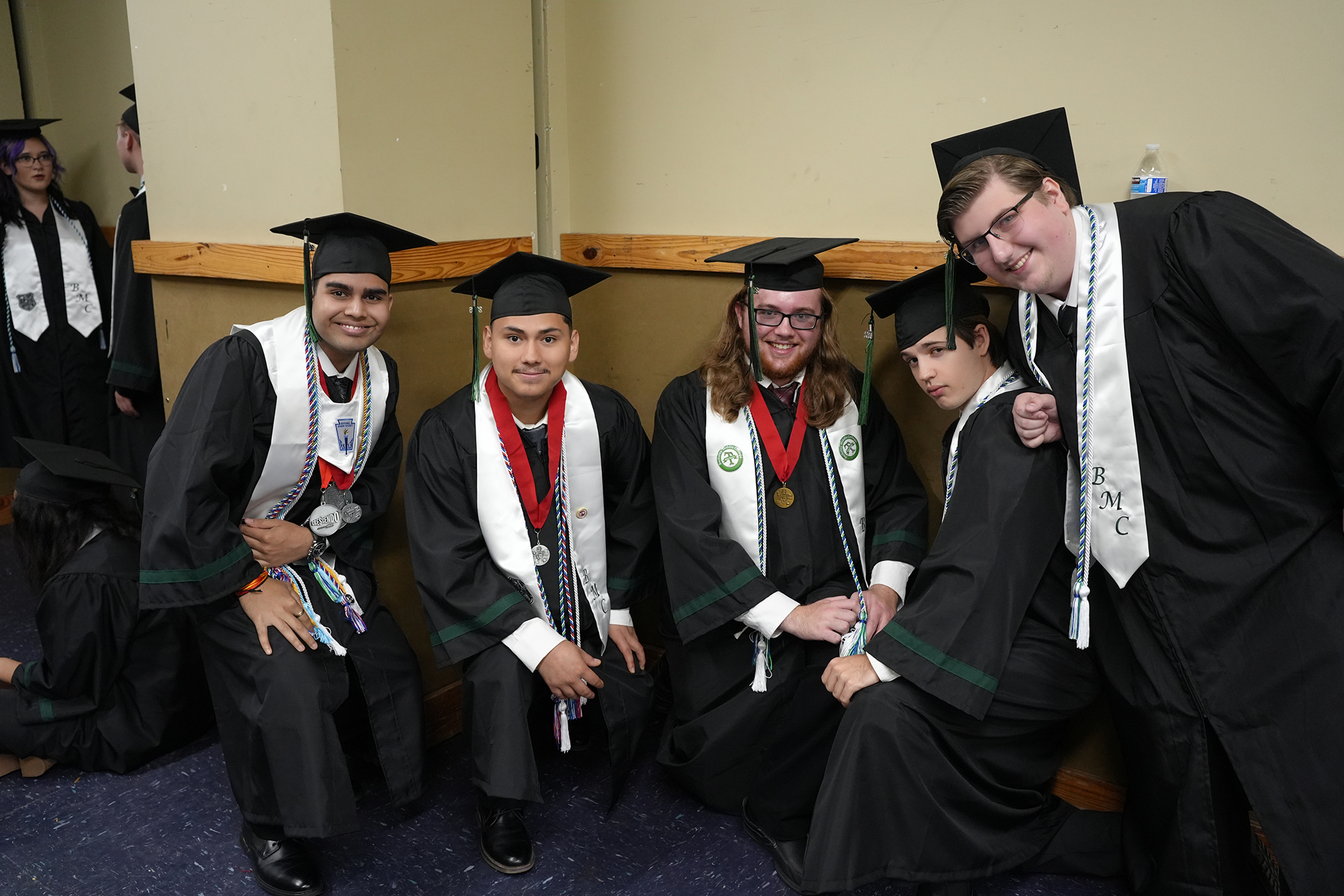 Five people in black gowns and mortarboards sit in a row, with one person standing behind them. They all wear medals and sashes.