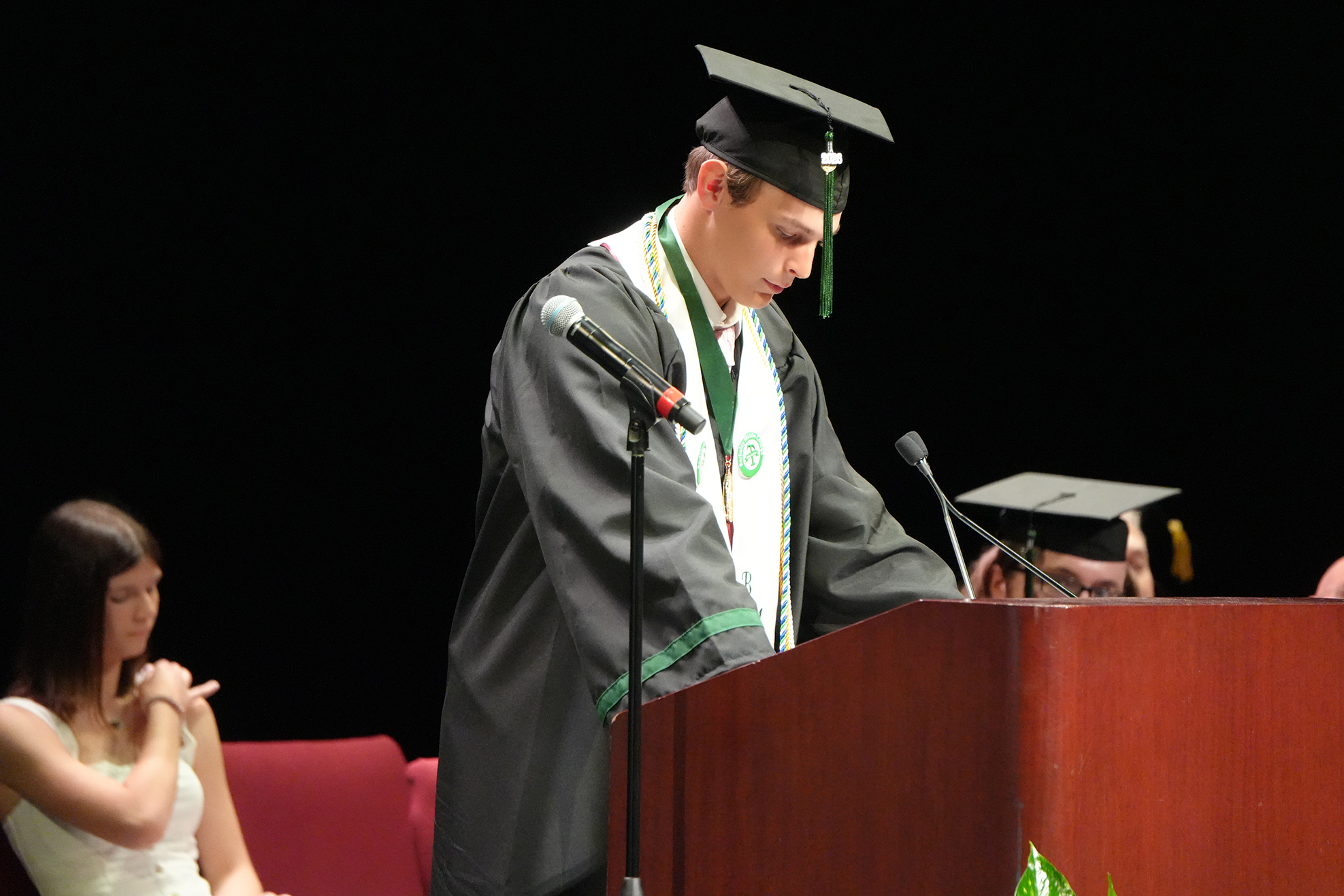 A man in a graduation gown and cap speaks at a podium. A woman in a white dress sits on a red chair behind him.