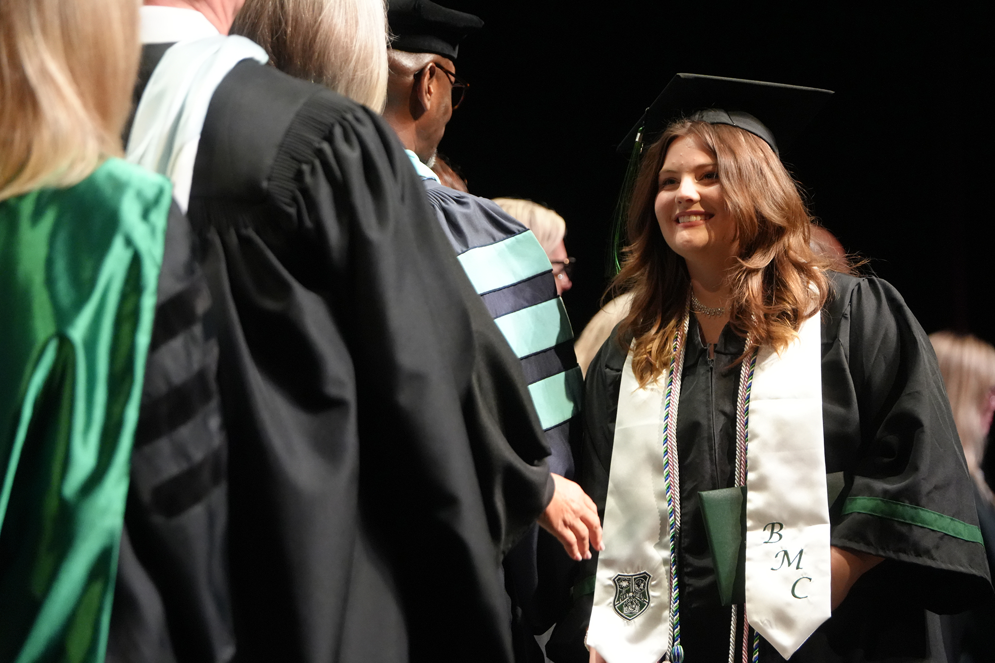 A graduate in a black gown smiles during a ceremony; others in black and green gowns are nearby.