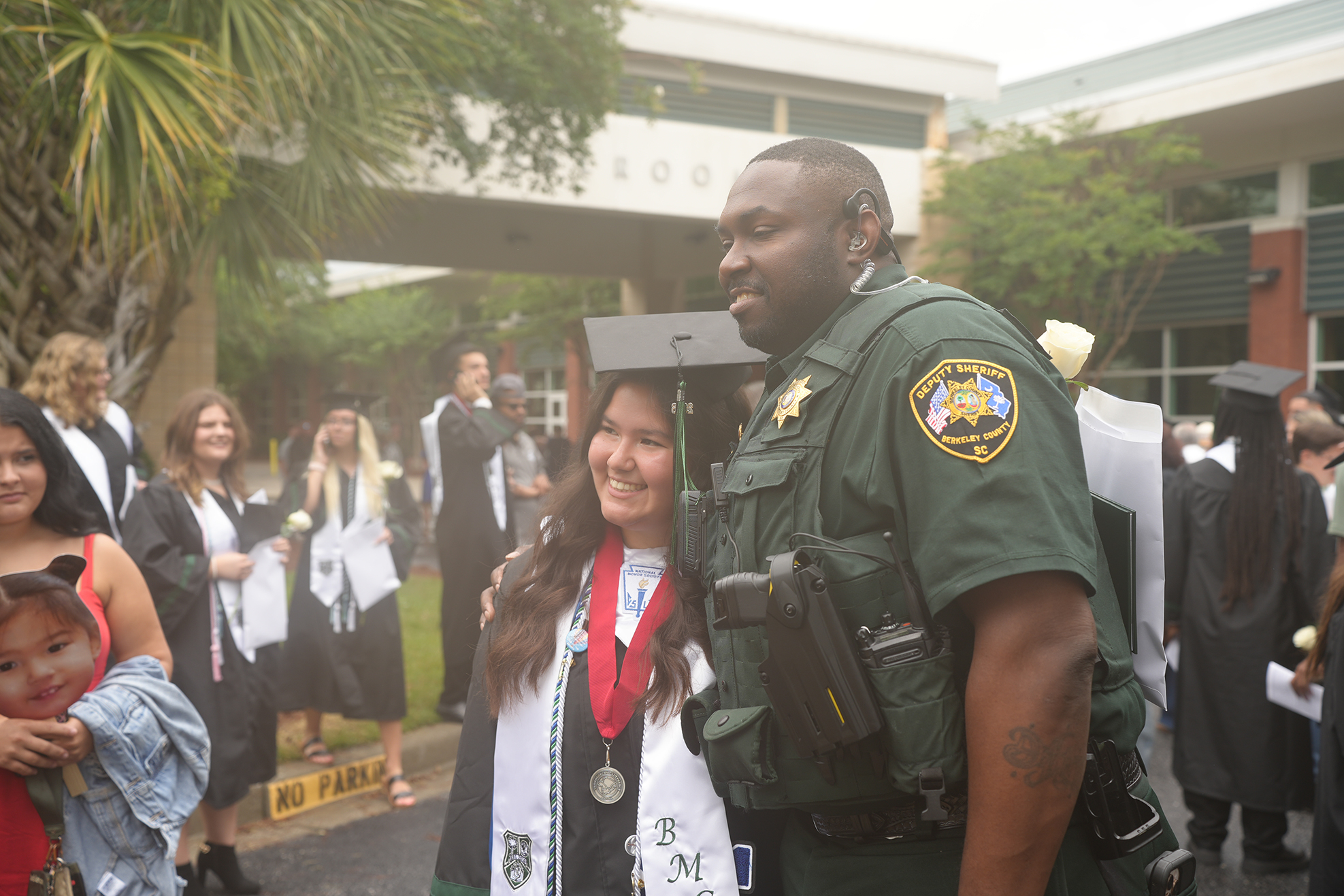 A police officer in green stands beside a woman in a white and blue graduation gown.