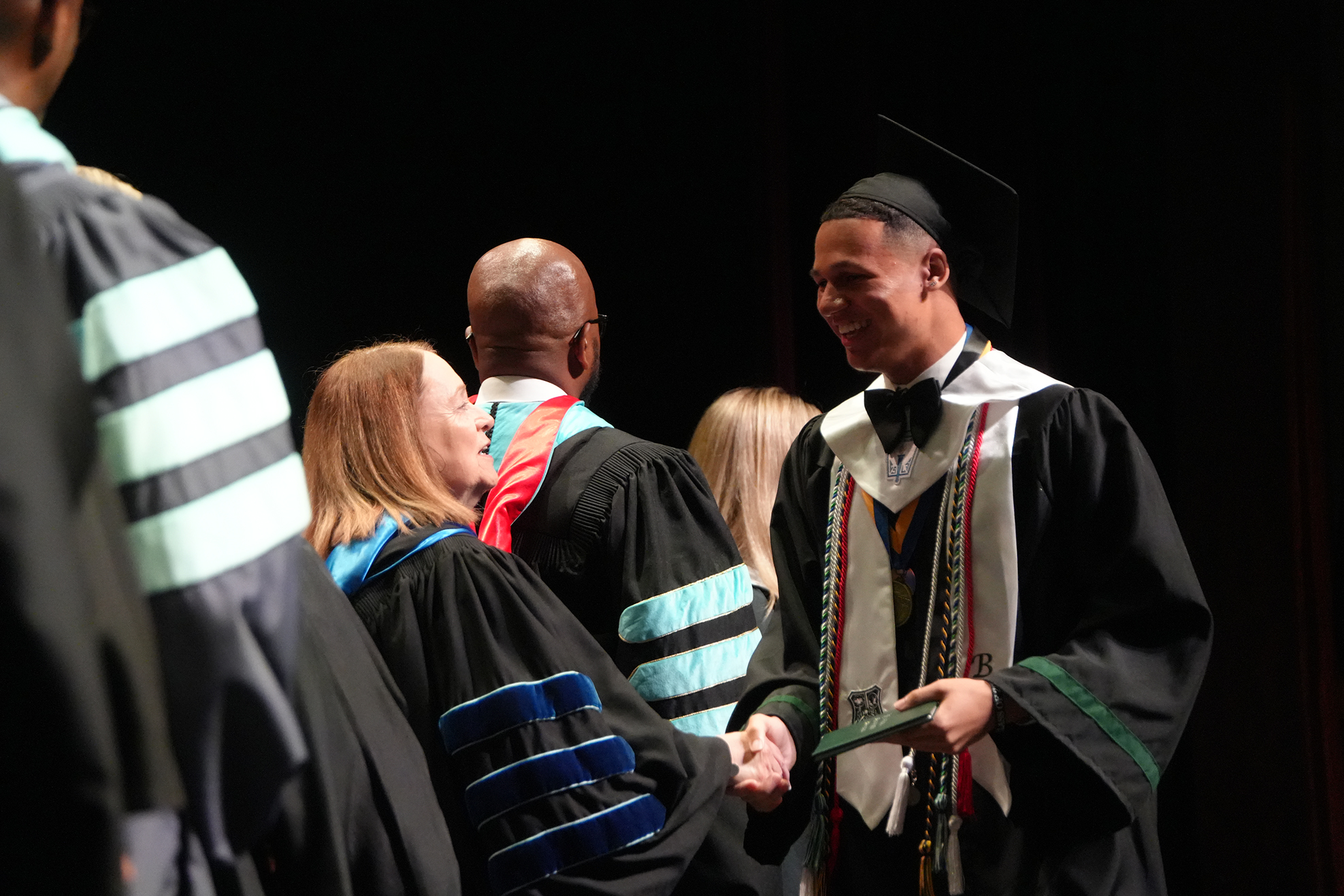 People in academic gowns and caps stand on a stage, possibly at a graduation. A man and woman shake hands.