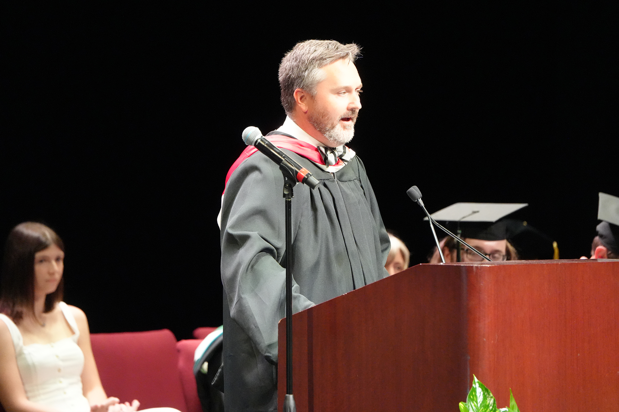 A man in a graduation gown speaks at a podium with a microphone, seated behind him is a woman in a white dress.