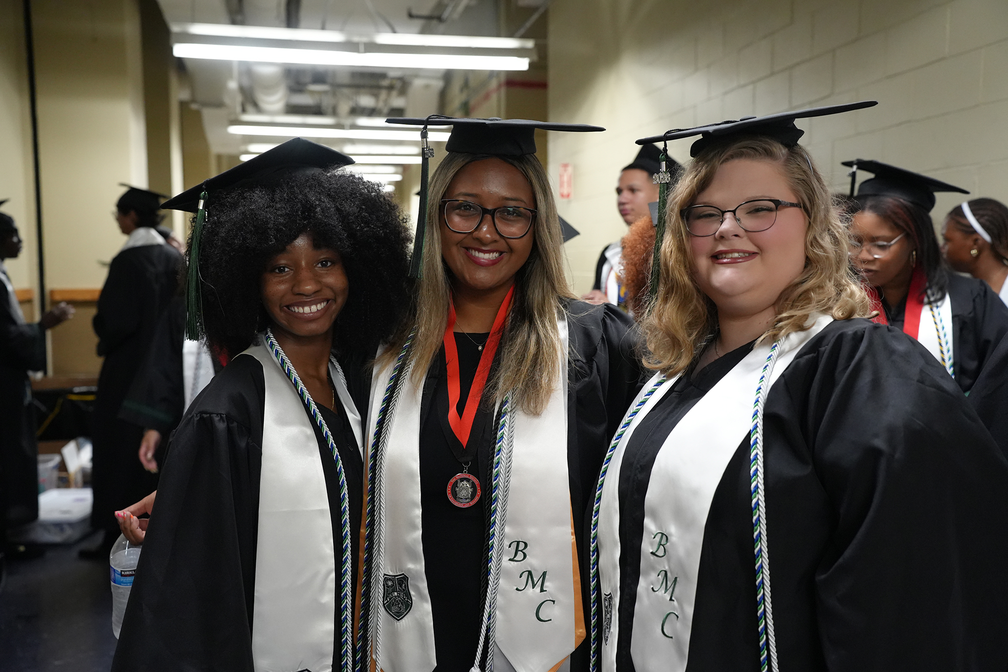 Three women in graduation robes and caps standing in a hallway. One wears a red ribbon, and two wear glasses.