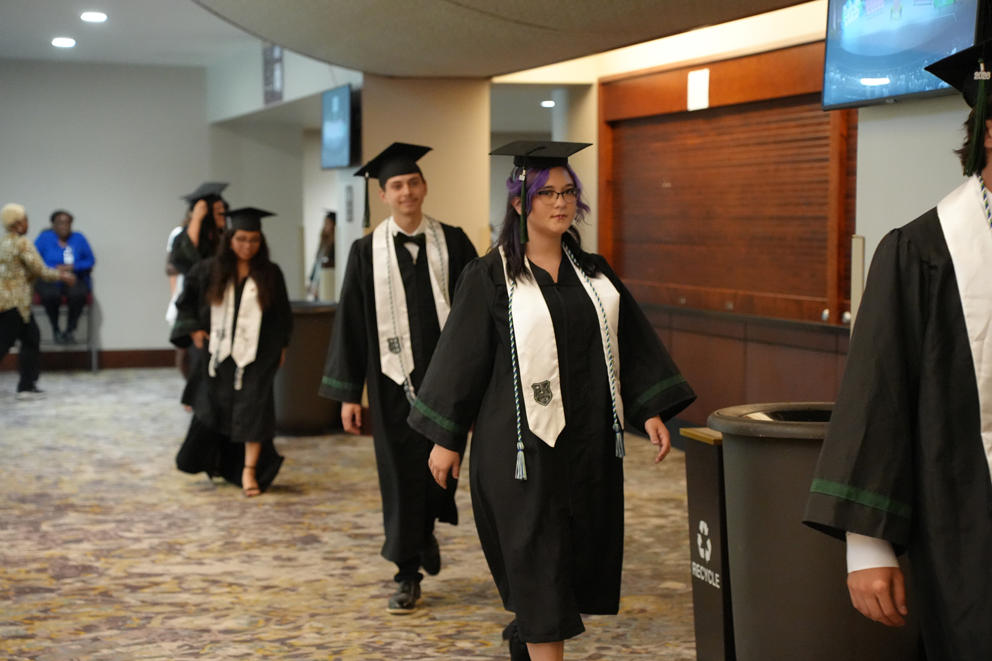 A group of students in black graduation gowns walk down a corridor with monitors and trash bins.
