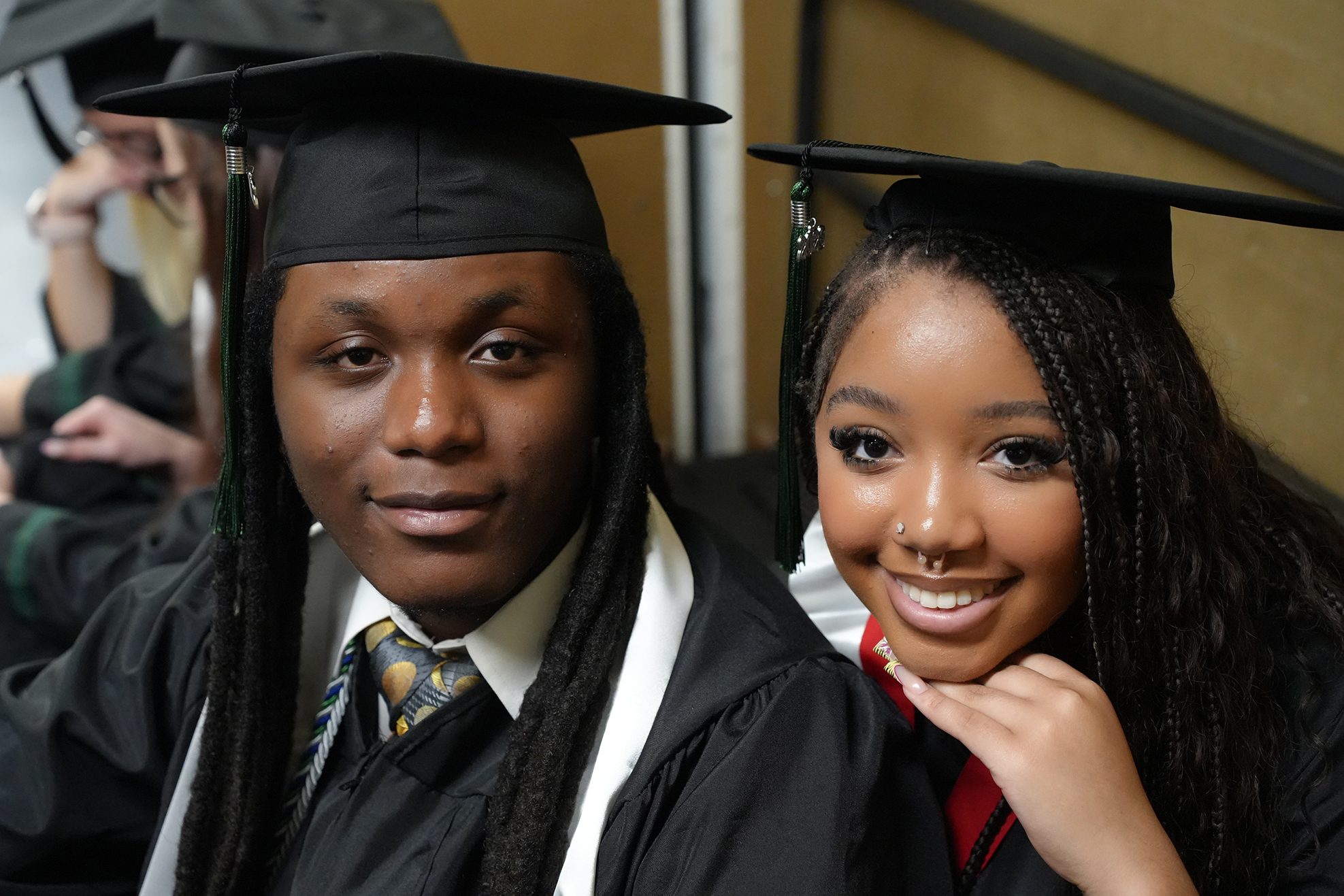 Two people in graduation robes and hats with tassels are sitting close together, smiling at the camera.
