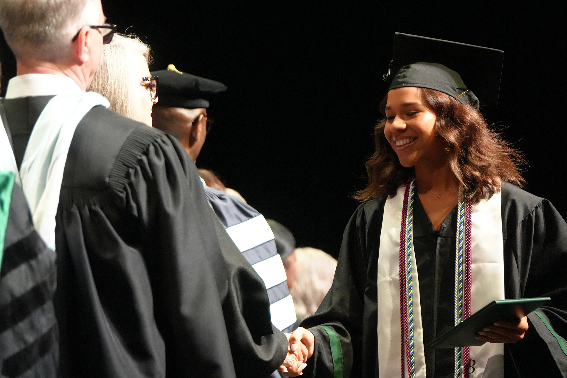 A graduate in a cap and gown smiles and shakes hands with a person in a gown.