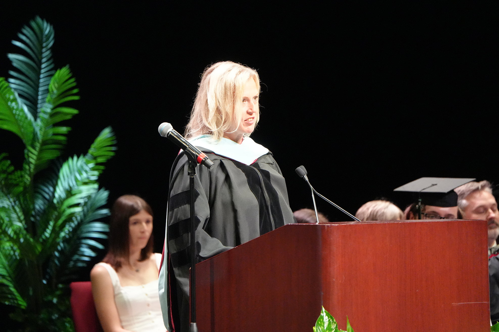 A person speaks at a podium, wearing a graduation gown. A plant and two microphones are in front.