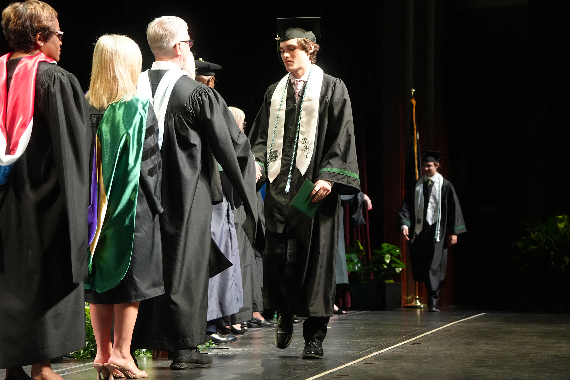 People in academic regalia walk on stage, one carrying a diploma. Behind them, a person stands, and a potted plant is visible.