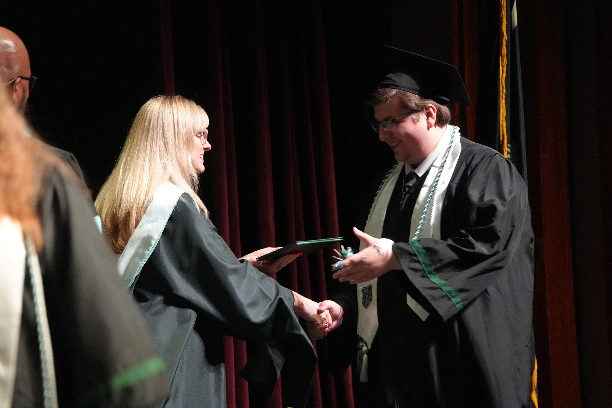 A man and a woman in graduation robes are shaking hands in front of a red curtain.