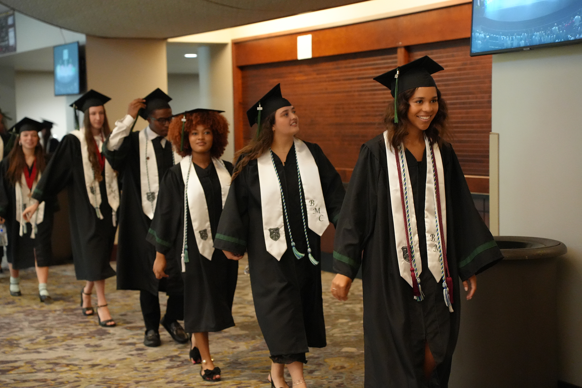 Graduates in gowns and caps walk down a hall, smiling and looking up, in a room with wooden walls and monitors.