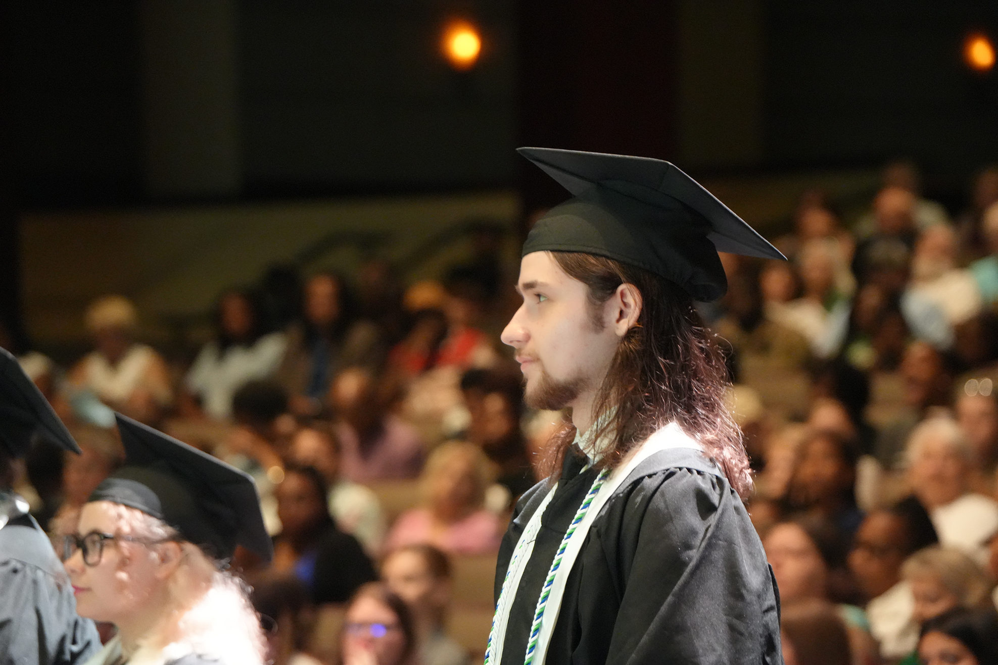 Graduates with long hair stand in a row, facing an audience at a ceremony. The background shows many people seated.